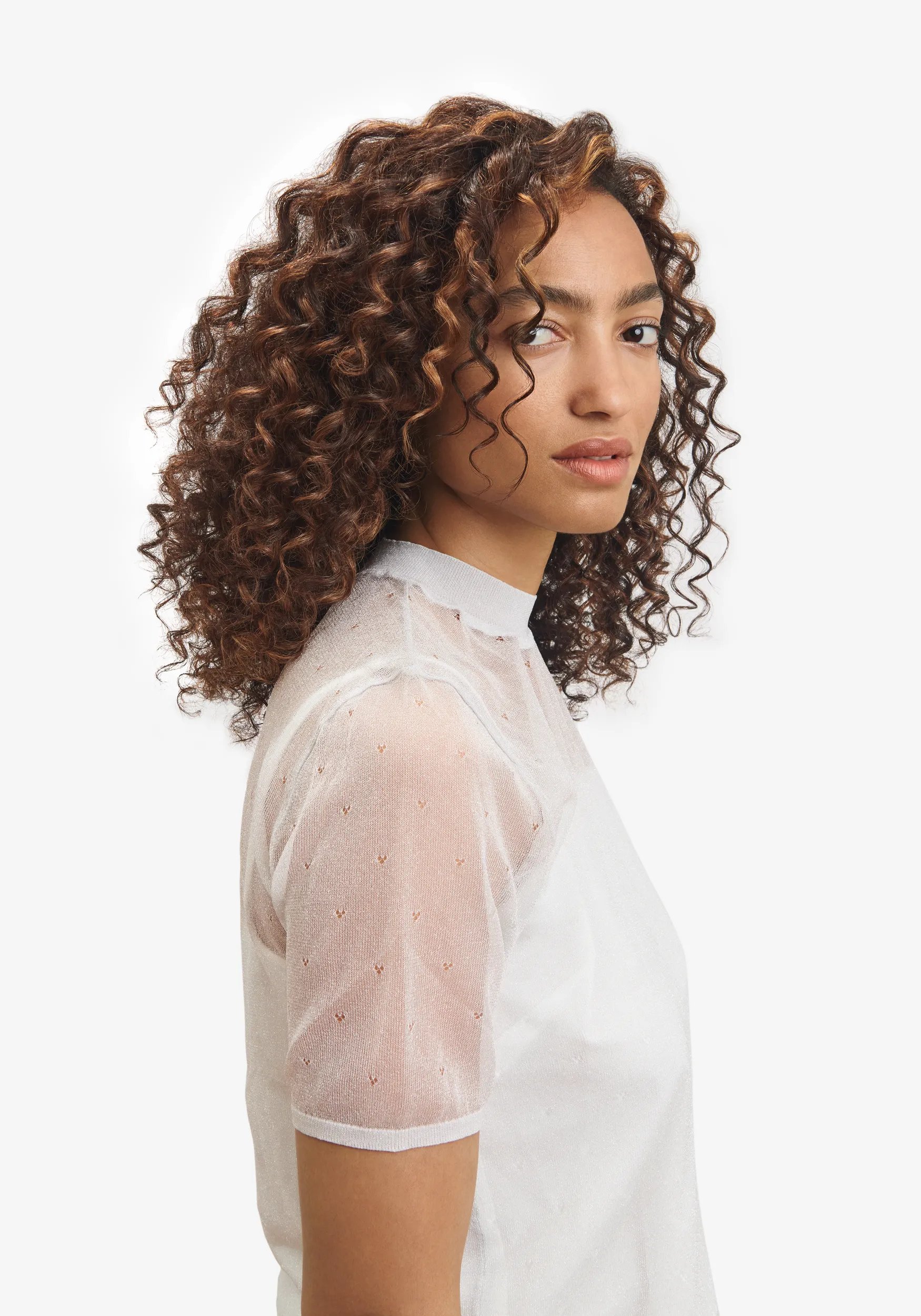 Side profile of model with brown shoulder length, curly hair. Wearing a white short sleeve shirt.