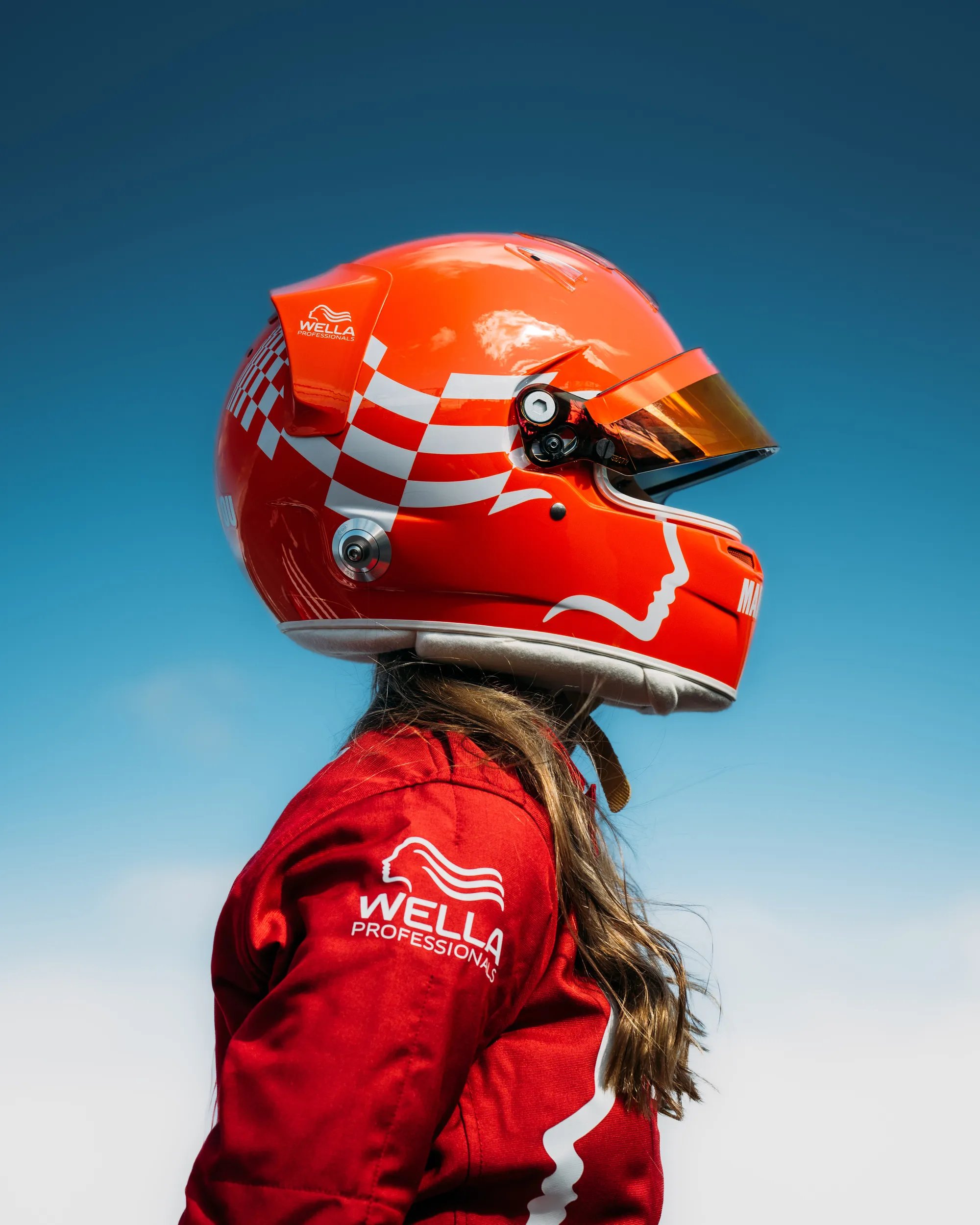 A red racing helmet displayed on a table alongside various Wella haircare and styling products, including bottles and tubes, with a flat iron and a black towel featuring the Wella logo.