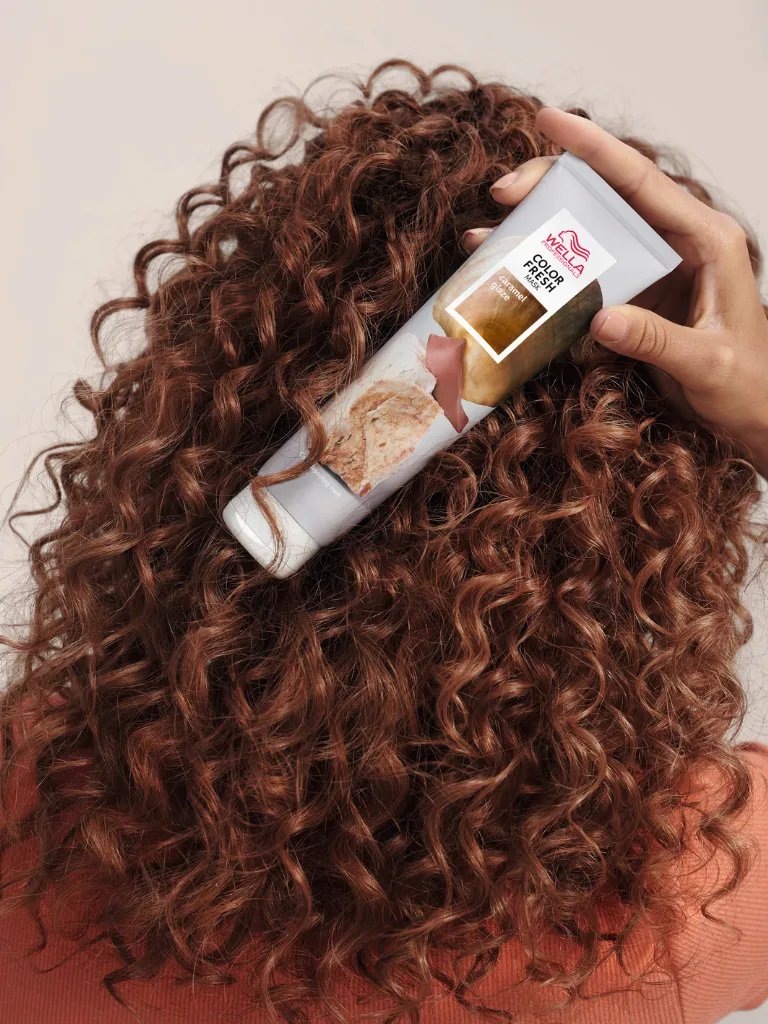 Model with curly, dark brown hair holds up a tube of Color Fresh Mask in Caramel Glaze.