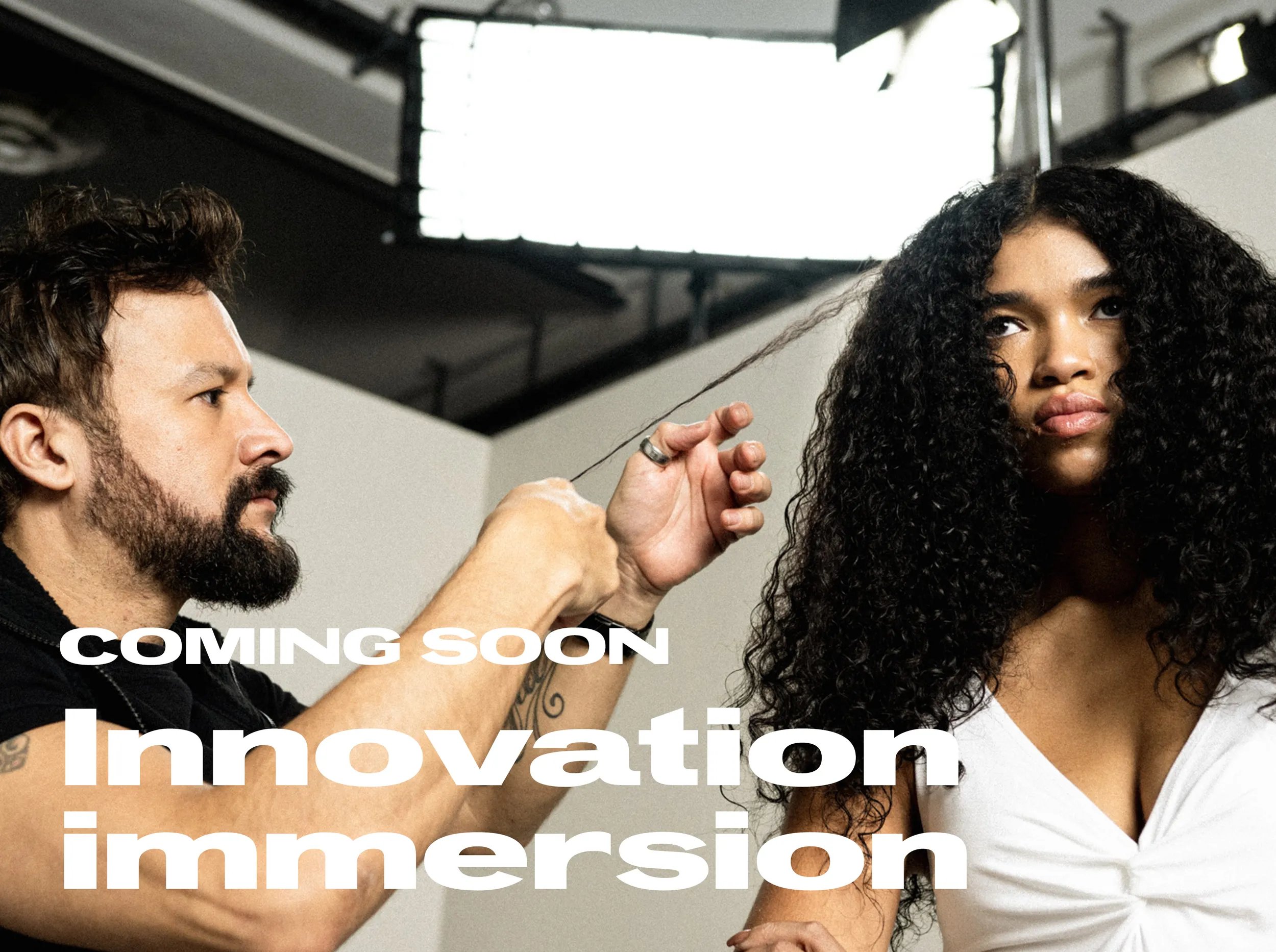 Immagine di un parrucchiere e di una modella con una bella chioma di capelli ricci. Lo stilista tiene in mano una ciocca di capelli della modella che guarda in avanti. Il testo in sovrimpressione recita “In arrivo Innovation Immersion"
