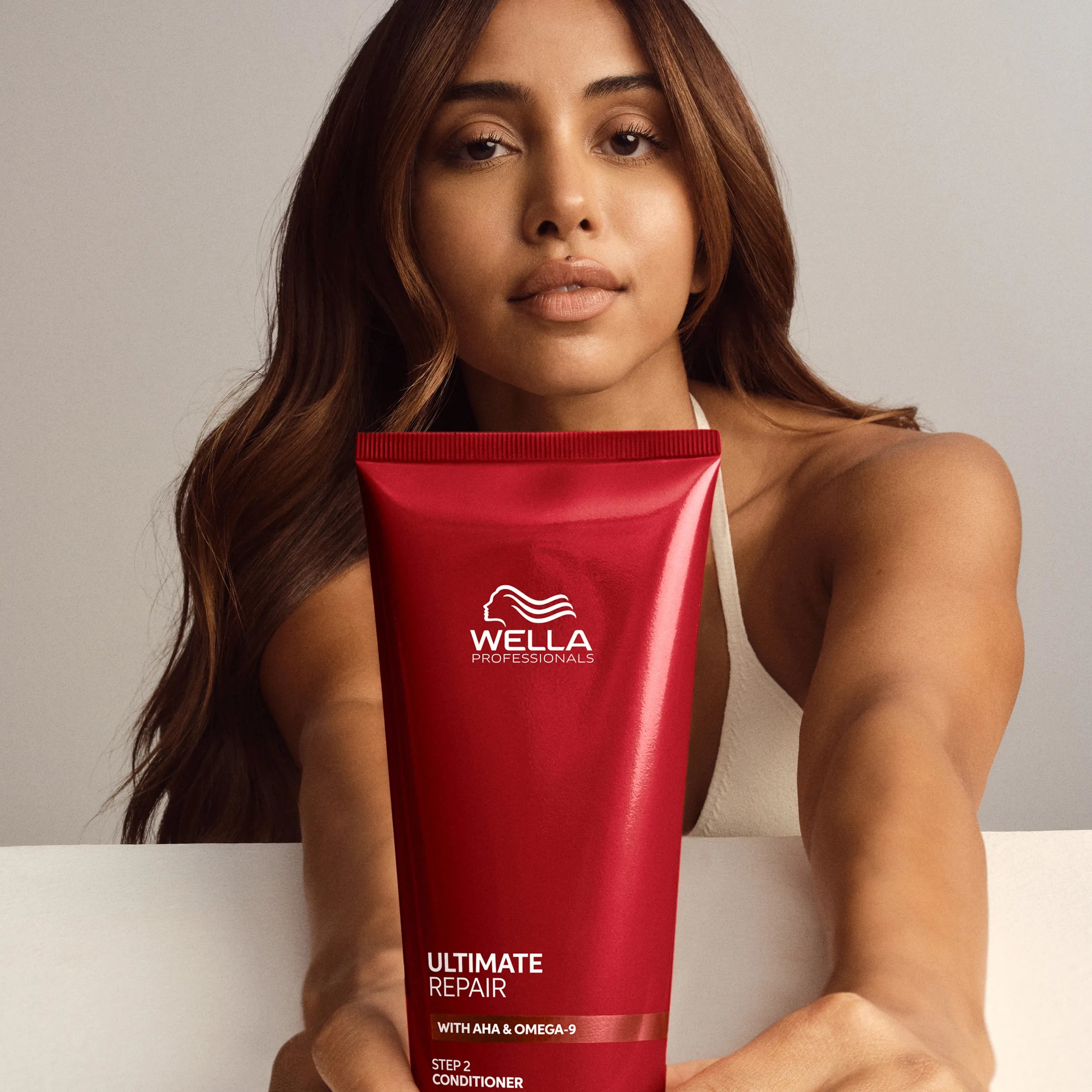 Woman with long, wavy, highlighted brown hair holding a red tube of Wella Professionals Ultimate Repair Step 2 Conditioner.