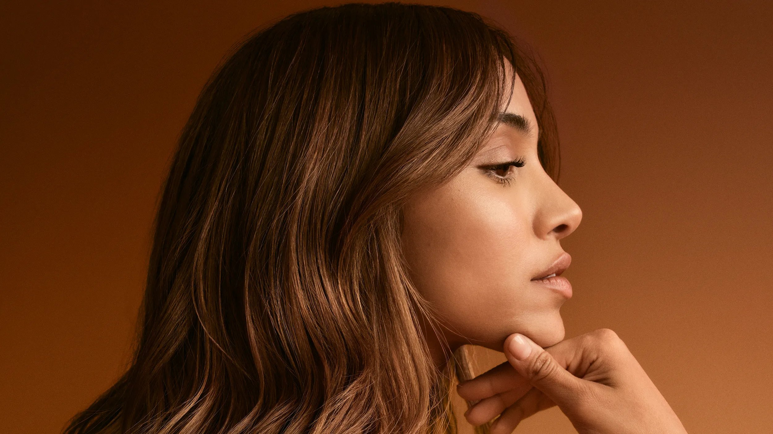 A side-profile portrait of a woman with long, light brown hair with her hand touching her chin. The background is a warm brown.
