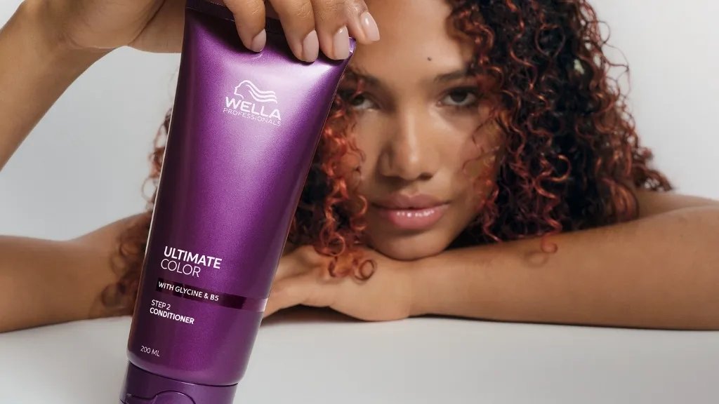 Model with curly, red-brown hair holds up a bottle of ULTIMATE COLOR Conditioner.
