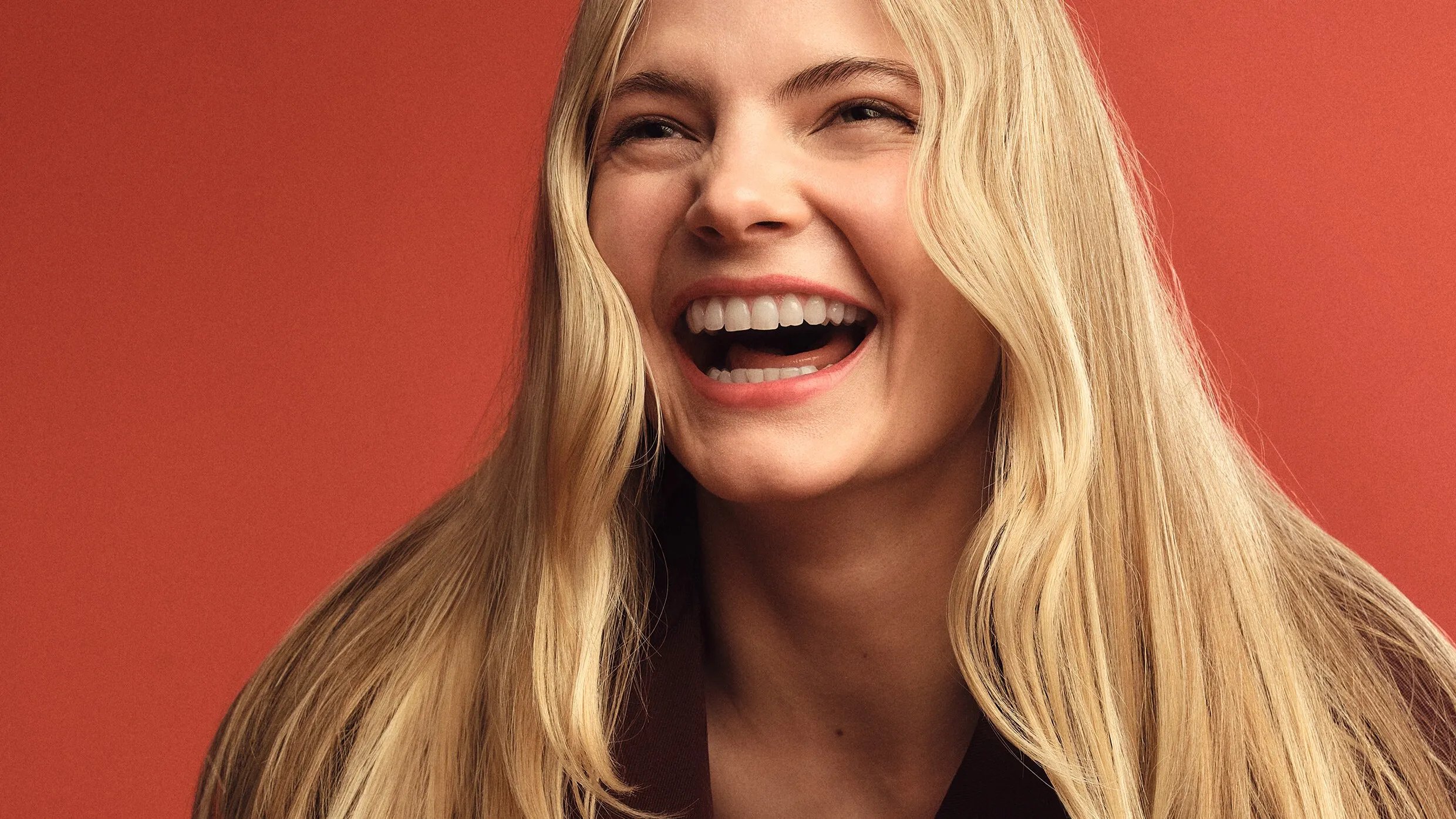 Model with long blonde hair, shown from the shoulders up against a warm red-orange background.