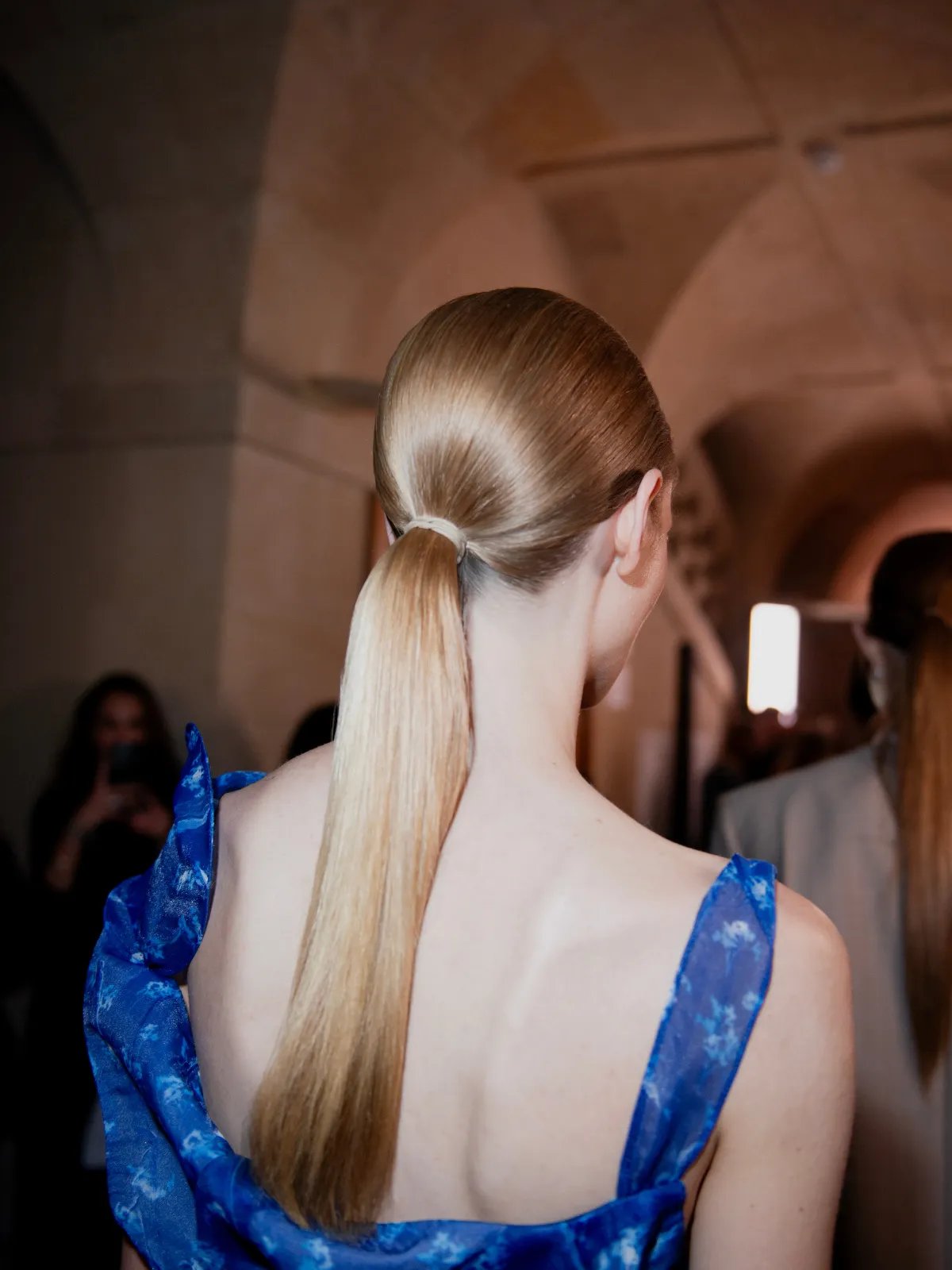 Back of model wearing a blue dress, with blonde hair slicked back in a low ponytail.