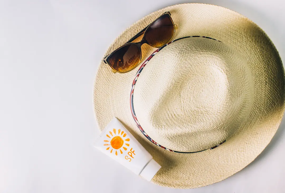 Flatlay photo of a beige straw sunhat, brown sunglasses and a tube of SPF lotion