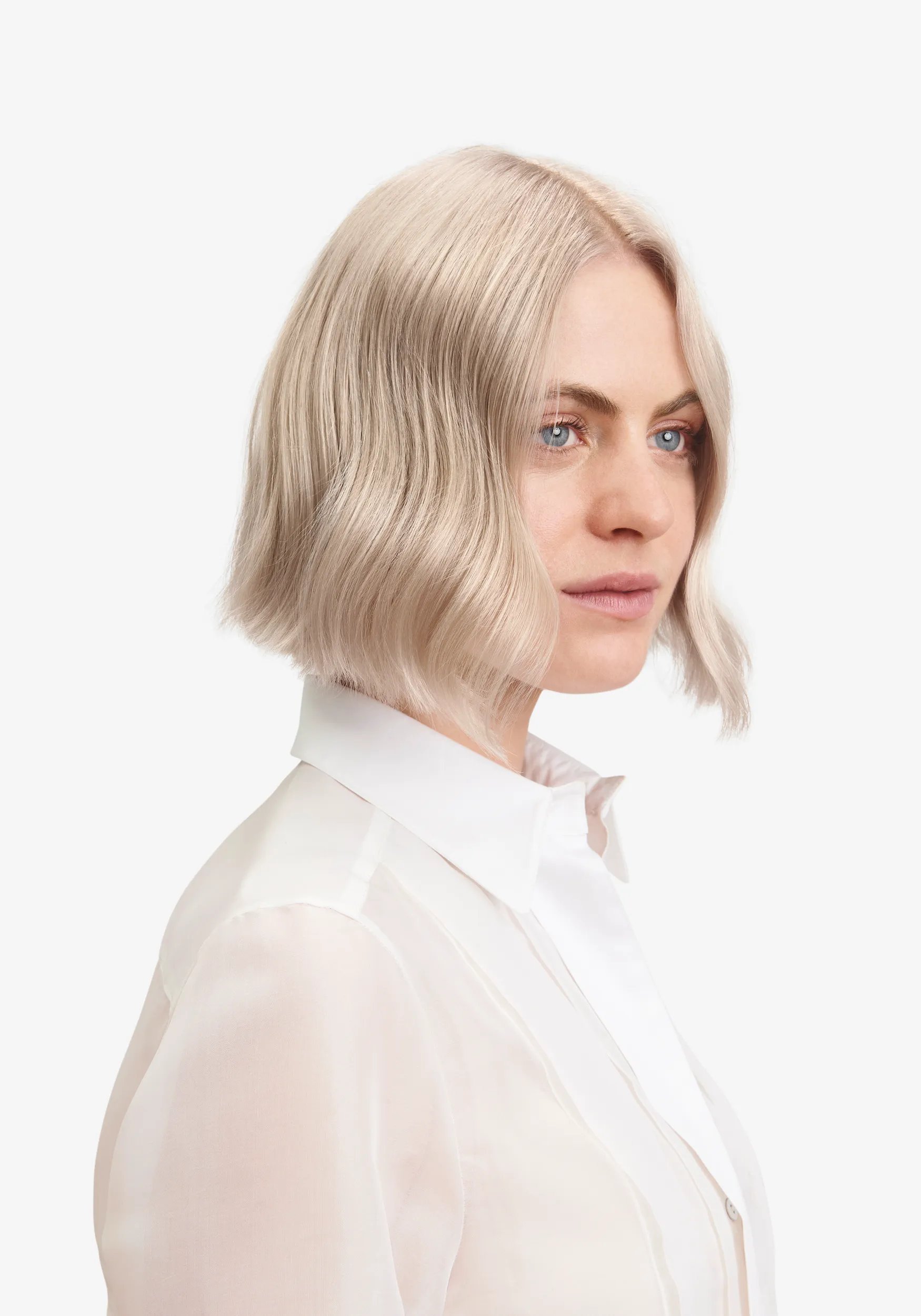 Profile view of a woman with a wavy, blonde bob hairstyle, wearing a sheer white collared shirt, against a white background.