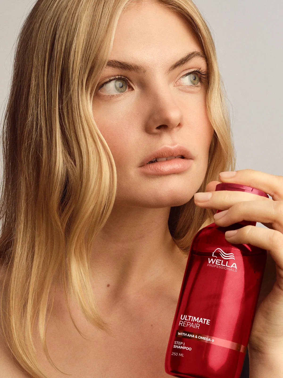 Model with long, tousled, blonde hair holds a bottle of ULTIMATE REPAIR Shampoo.