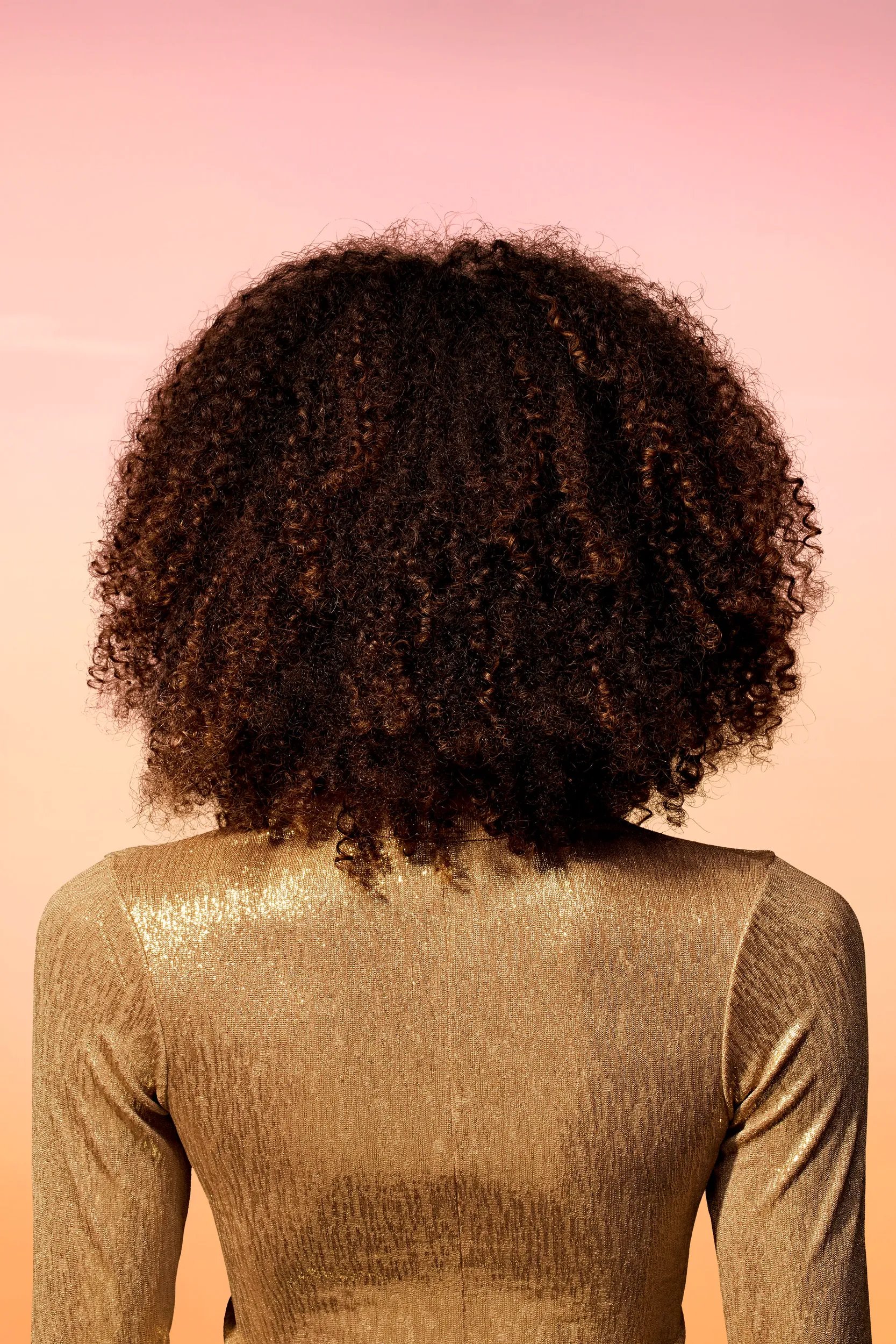 A model with a dark brown, shoulder length, layered afro and golden brown highlights faces away from the camera. Curls are not well defined.
