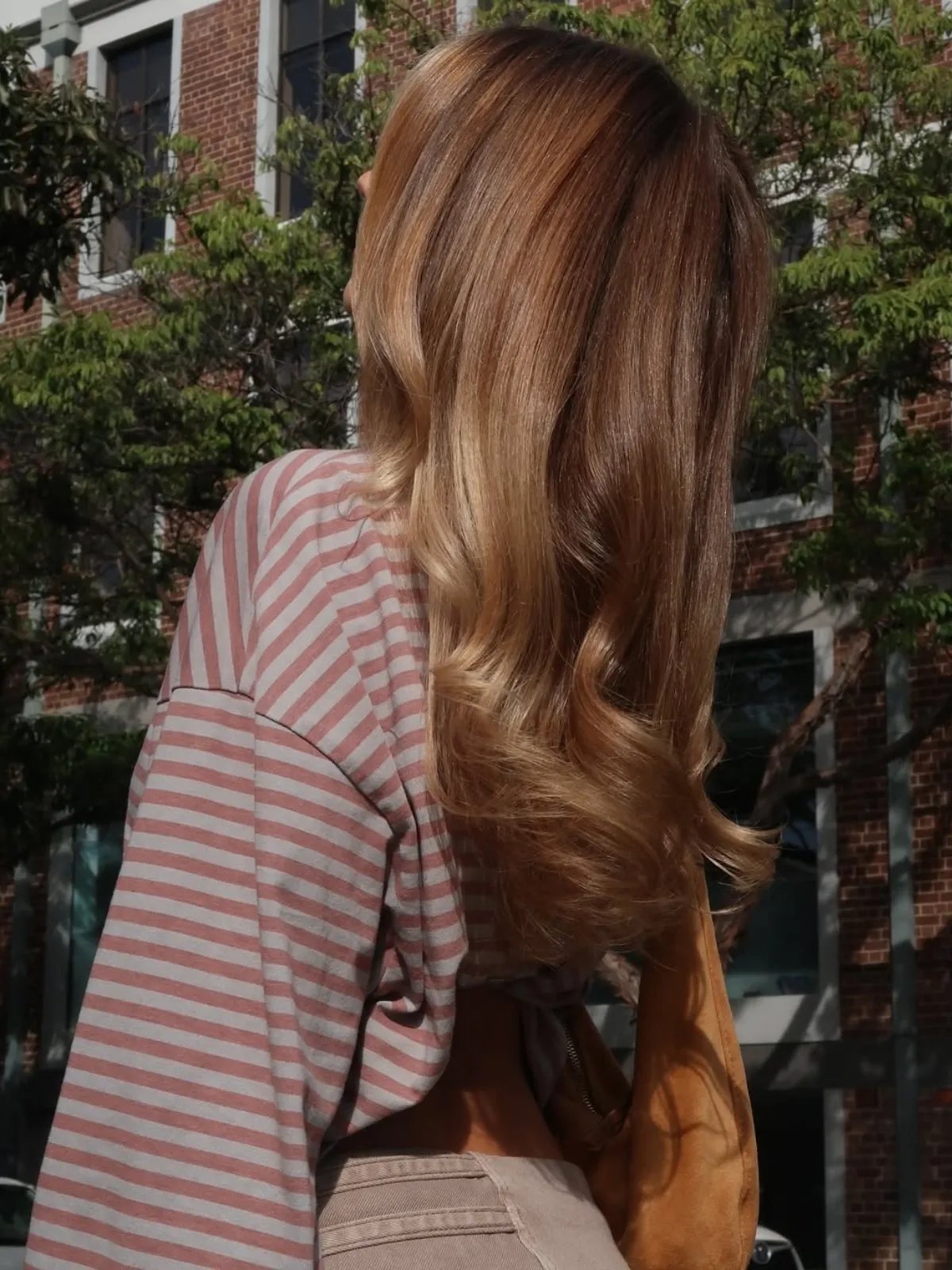 Model with long, loosely curled, light brown hair and subtle blonde accents through the ends.