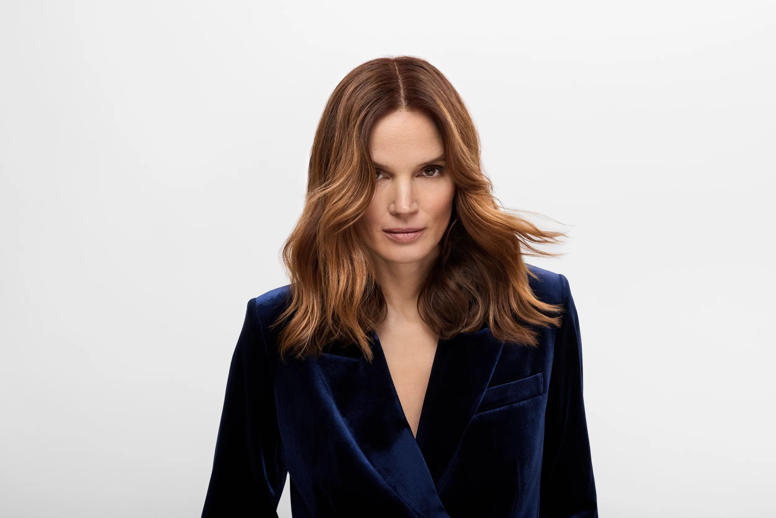 Close-up portrait of a model with shoulder-length, wavy copper-brown hair against a light grey background.
