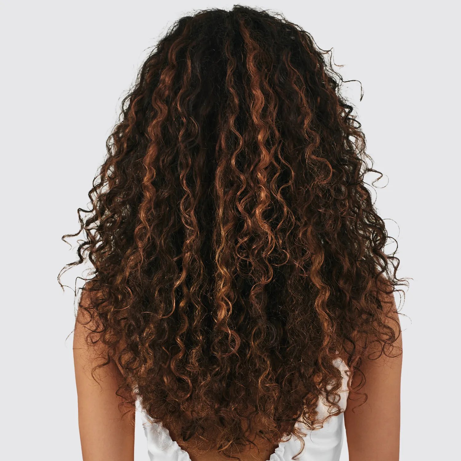Image showing the back of a woman with long, dark, curly hair with subtle blonde highlights, wearing a white top against a neutral background.