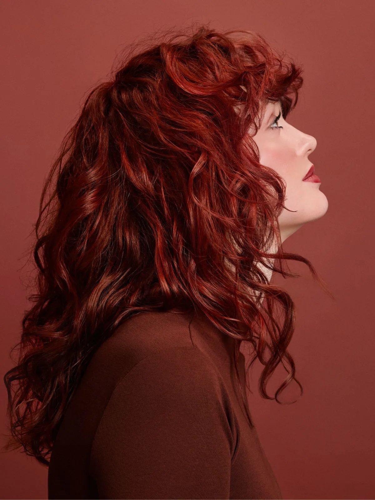 Model with shoulder-length auburn curly hair and bangs, wearing a brown leather jacket over a high-neck top. The individual is centered against a warm terracotta background, looking directly at the camera. The image features soft lighting and a monochromatic palette of reddish-brown tones.