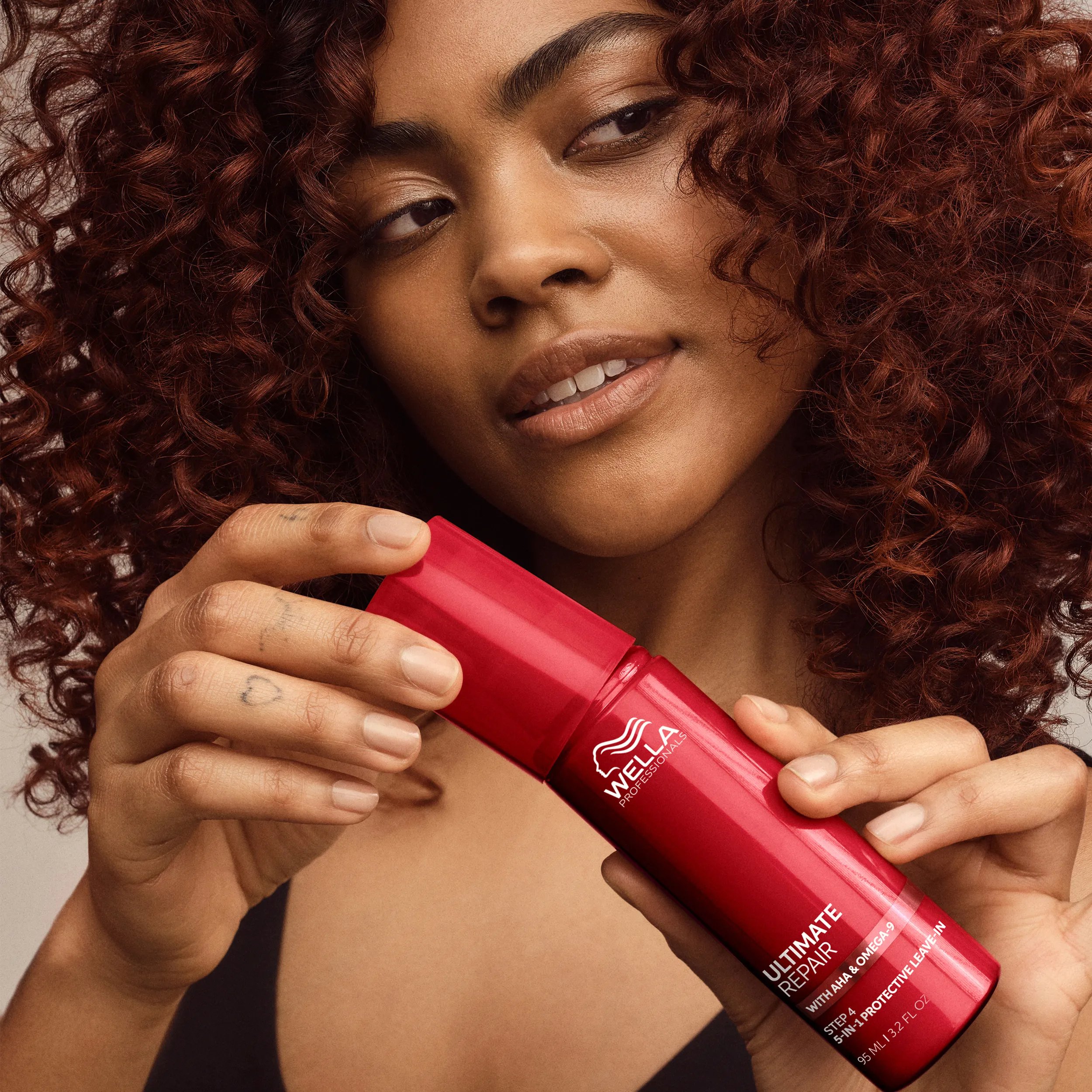 Woman with curly auburn hair holding and displaying a red bottle of Wella Professionals Ultimate Repair Step 4 5-in-1 Protective Leave-In Treatment.
