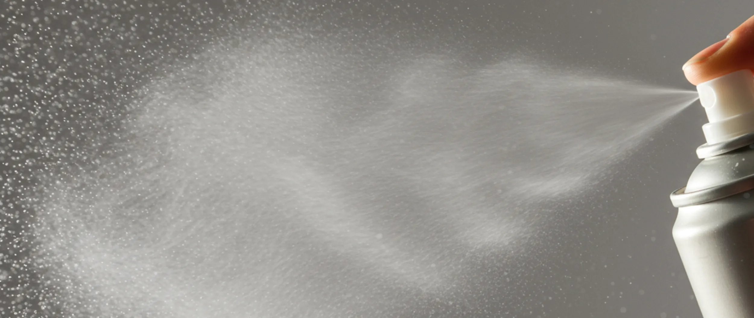 A hand presses the nozzle of an aerosol spray can, releasing a fine mist against a gray background.