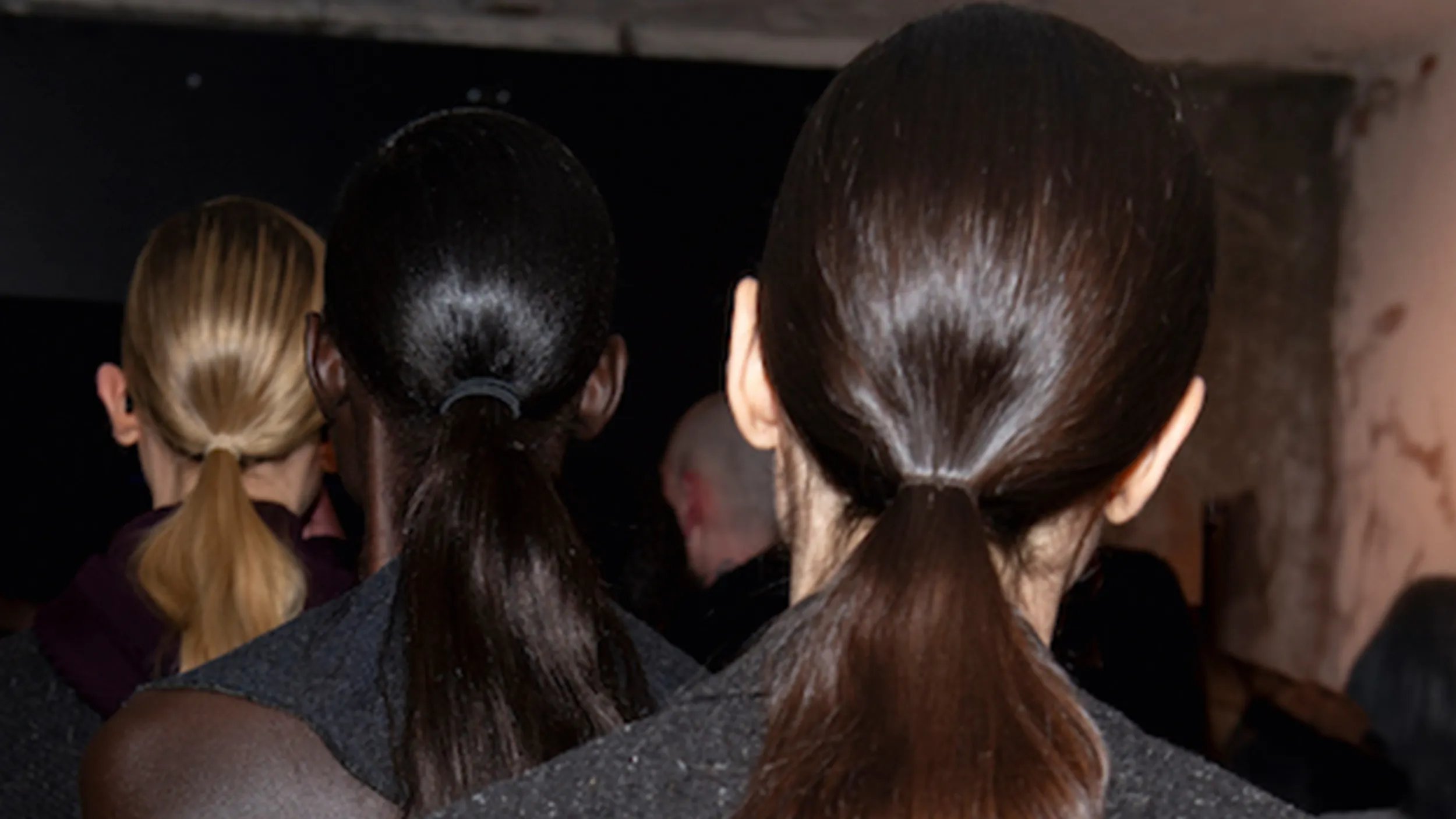 Back view of several models with sleek ponytails, showcasing different hair colors and textures backstage.