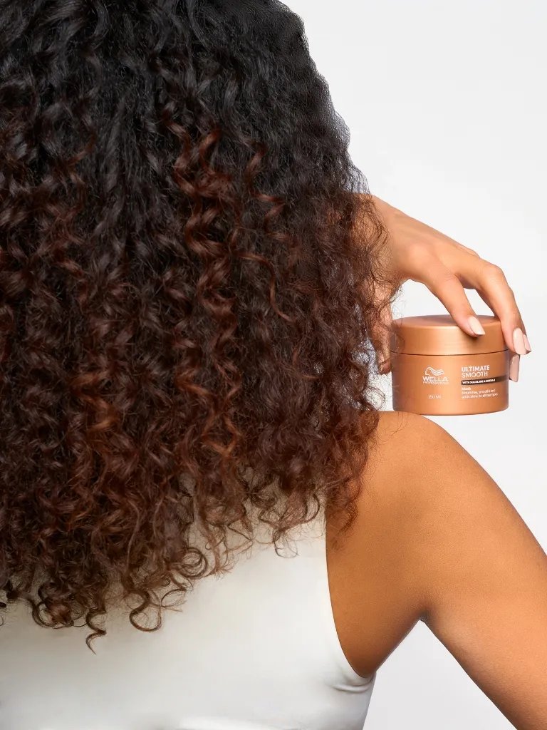 Model with dark brown, curly hair holds a tub of ULTIMATE SMOOTH Intense Mask.