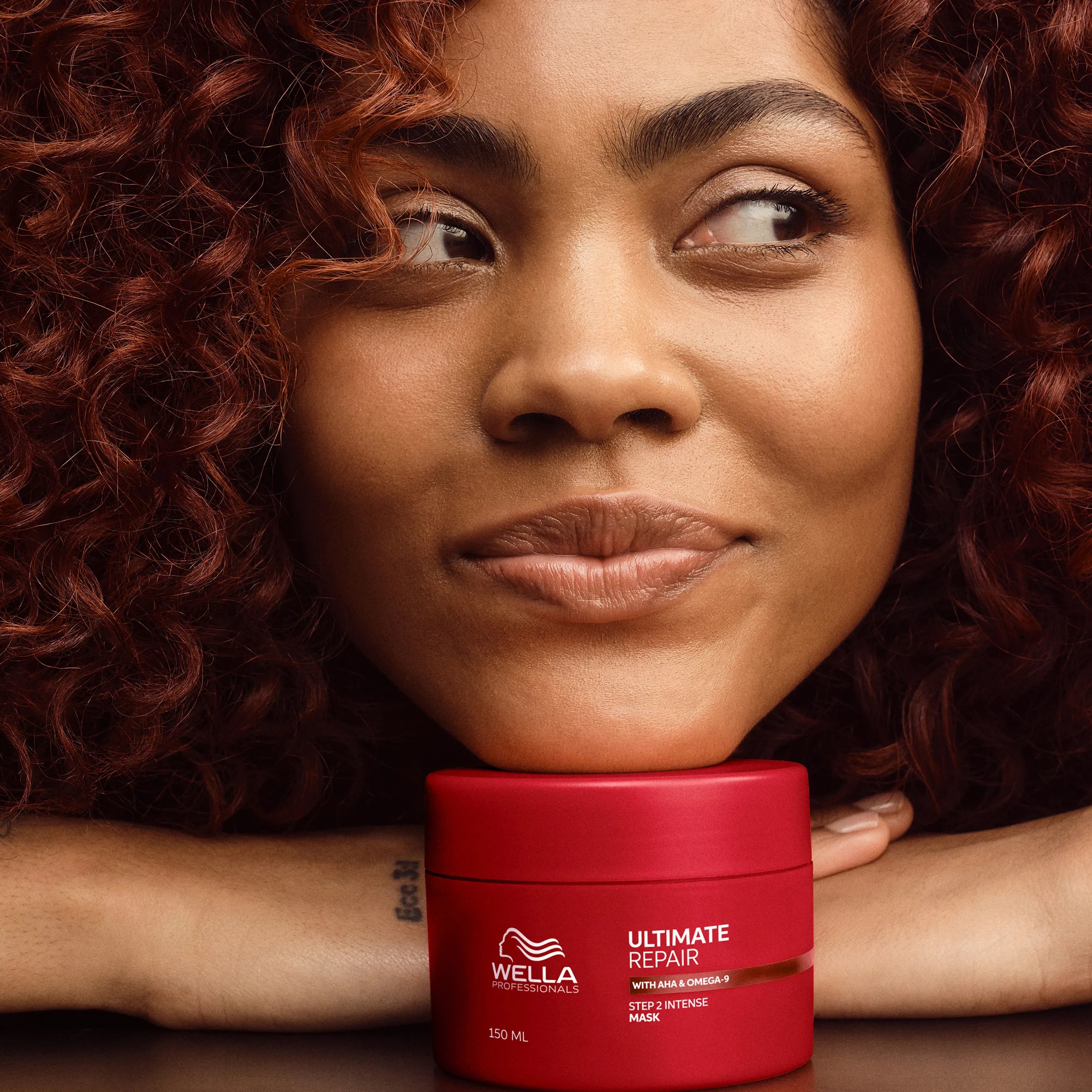 A smiling woman with curly auburn hair rests her chin on a red jar of Wella Professionals Ultimate Repair Step 2 Intense Mask.