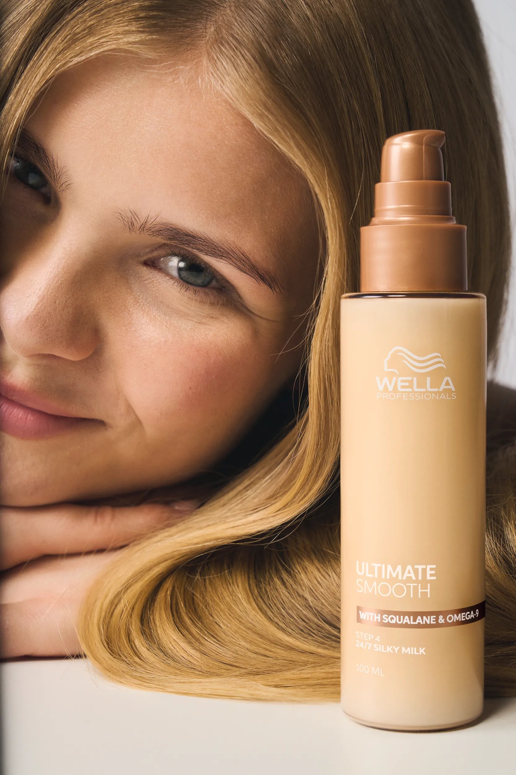 Bote de Wella Professionals ULTIMATE SMOOTH 24/7 Silky Milk en primer plano con un acercamiento de un cabello de apariencia suave al fondo, con el texto: “Fórmula ligera para retoques diarios en cualquier lugar”.