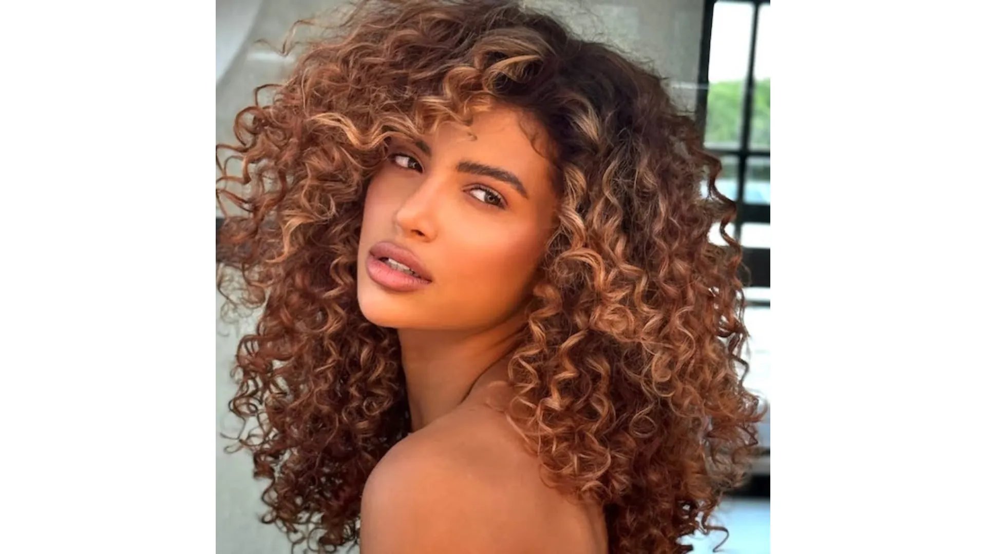 Model with long, dark brown, curly hair featuring golden, sun-kissed highlights.