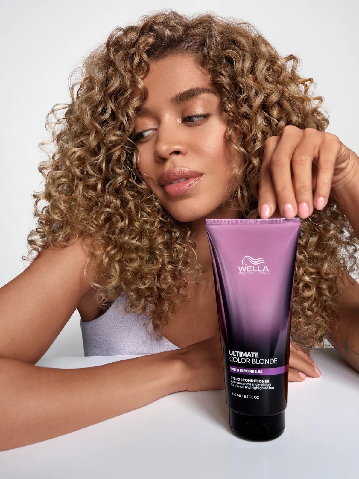 Model with brown-blonde, curly hair holds a bottle of ULTIMATE COLOR BLONDE Conditioner.