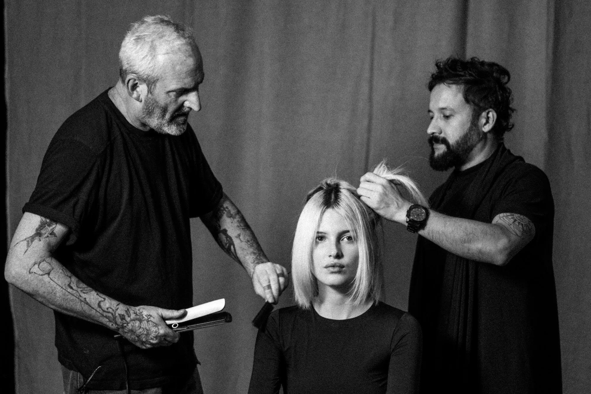 Black and white image of a model seated between two hairstylists. One hairstylist is holding straighteners and a comb and the other adjusting the model's hair