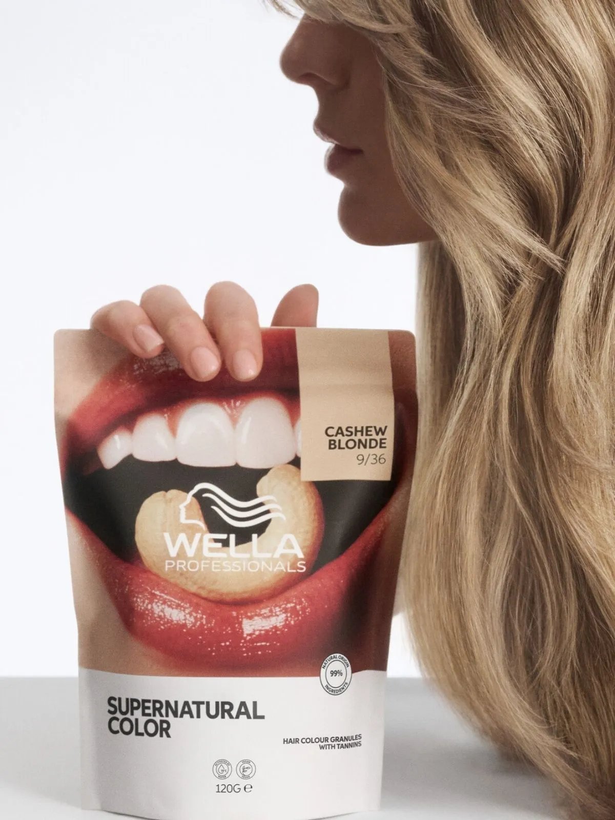 Model with long, tousled, blonde hair holding a pouch of Supernatural Color in Cashew Blonde.