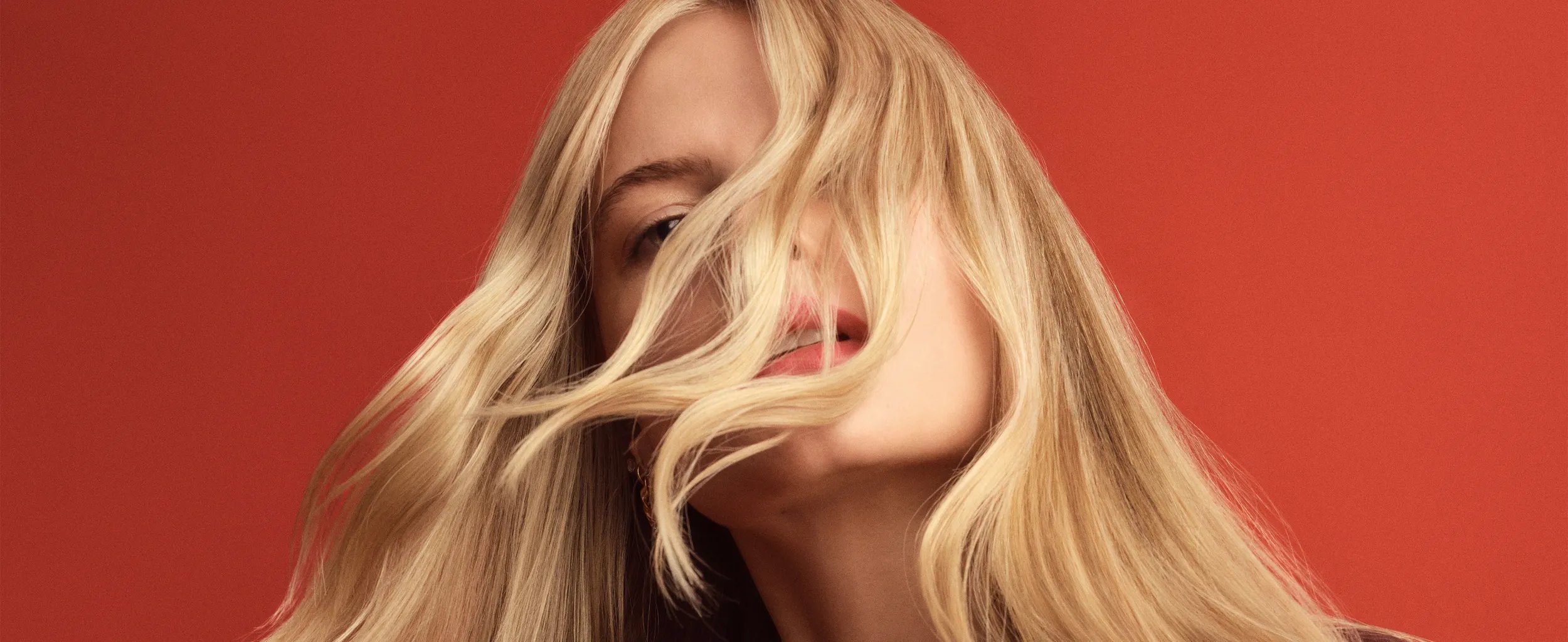 Blonde woman with long hair blowing in the wind, wearing a burgundy blazer against a red background.
