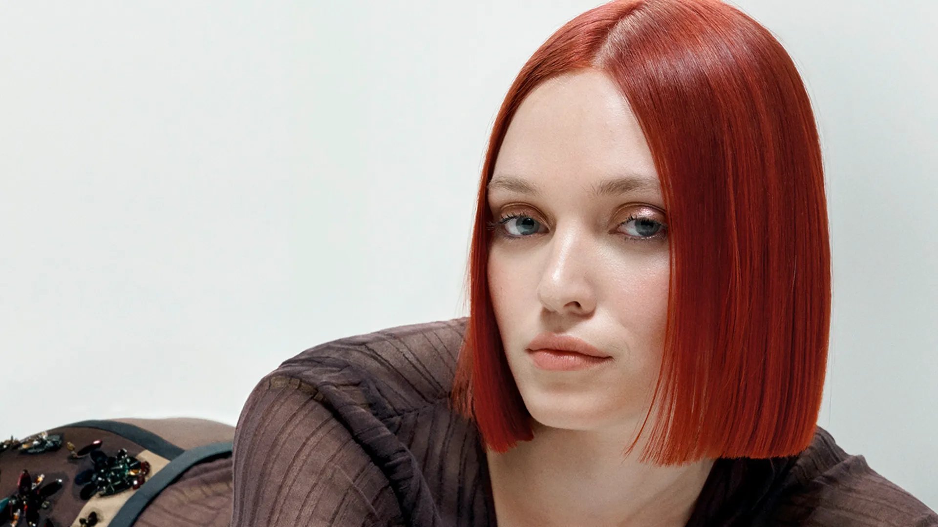 Image of model with short, straight and red hair laying down and looking forward and smiling slightly. Model is wearing a brown outfit. 