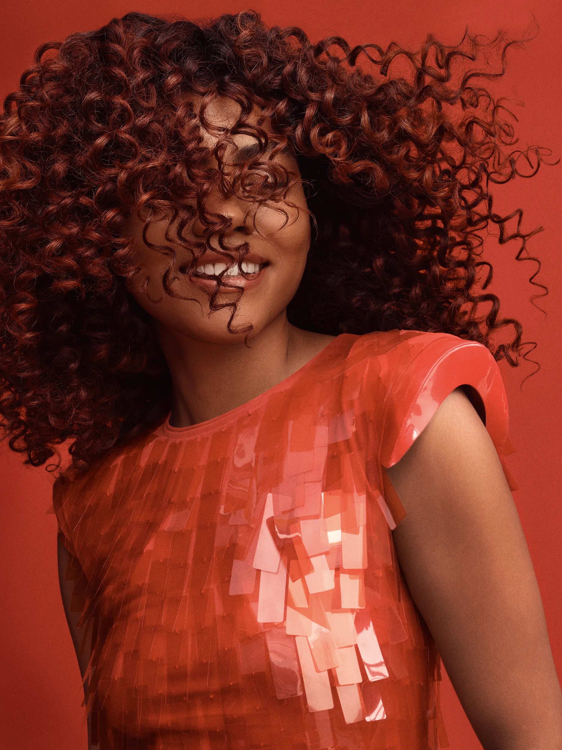 Woman smiling with curly red hair partially covering her face, wearing a sheer red top against a matching background.