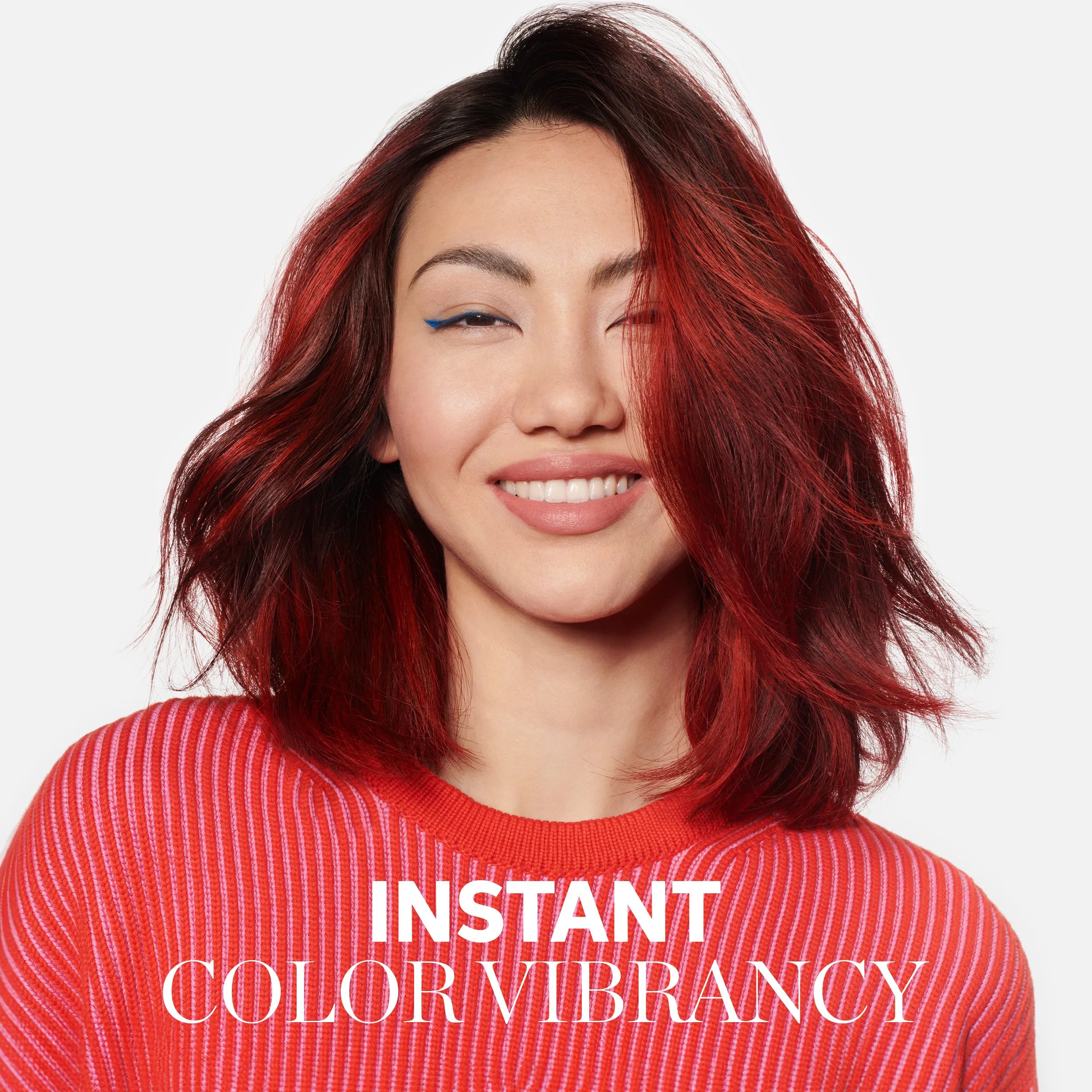 Photograph of a young woman with voluminous shoulder-length red hair. She is smiling and wears a striped red jumper and blue winged eyeliner. Overlaid text reads 'instant color vibrancy'.