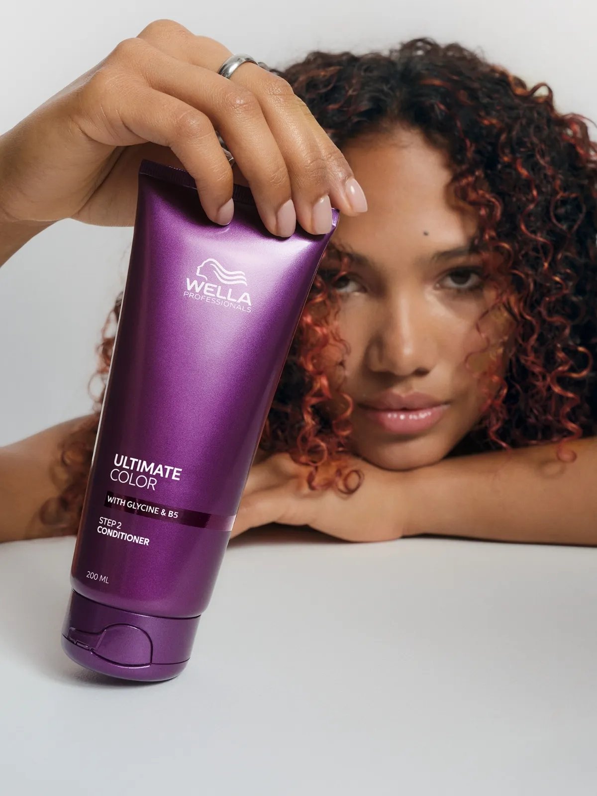 Model with red-brown, curly hair holds a bottle of ULTIMATE COLOR Conditioner.