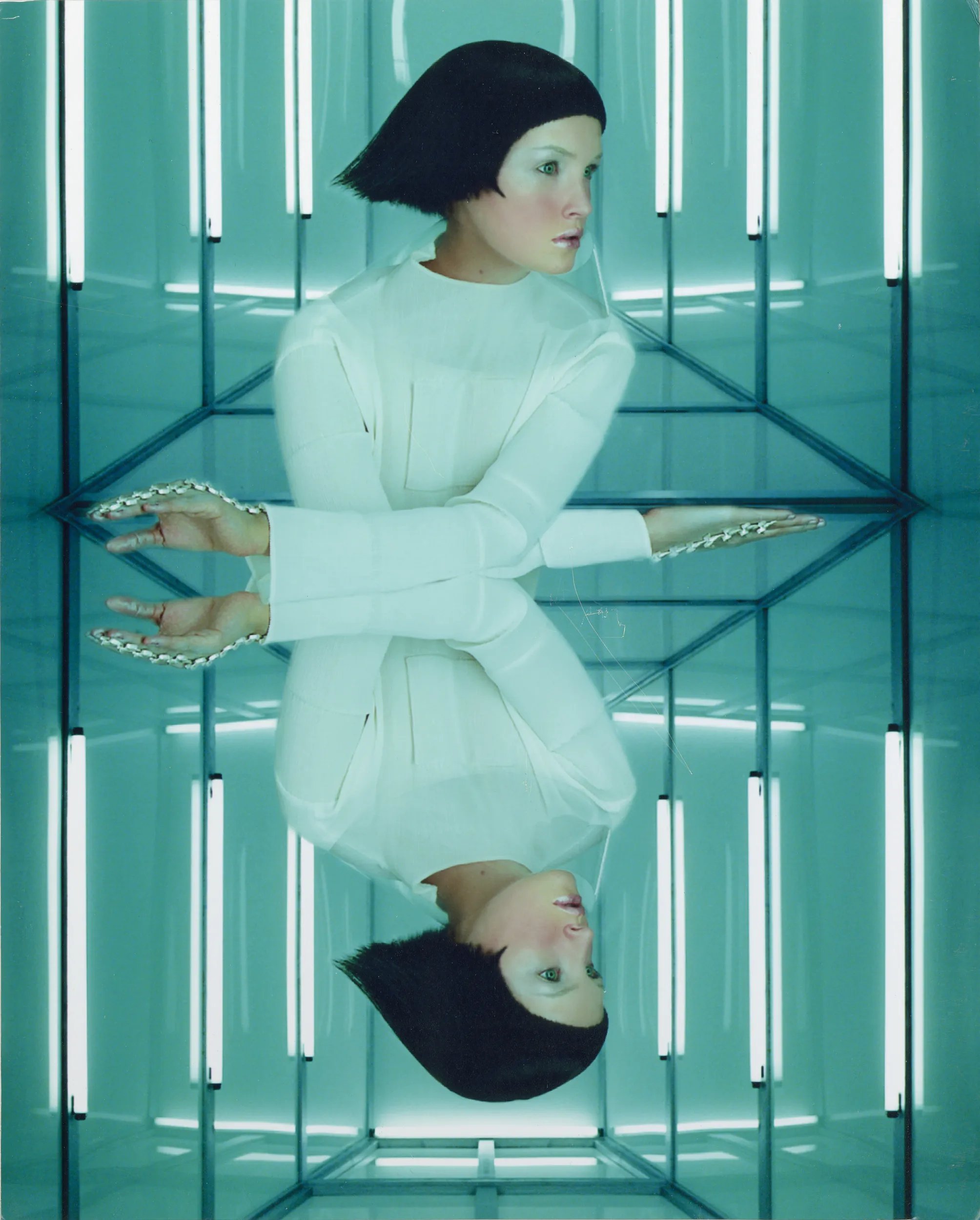 A model in a white outfit with metallic make up and short black hair reflected in a glossy surface, surrounded by linear lights.