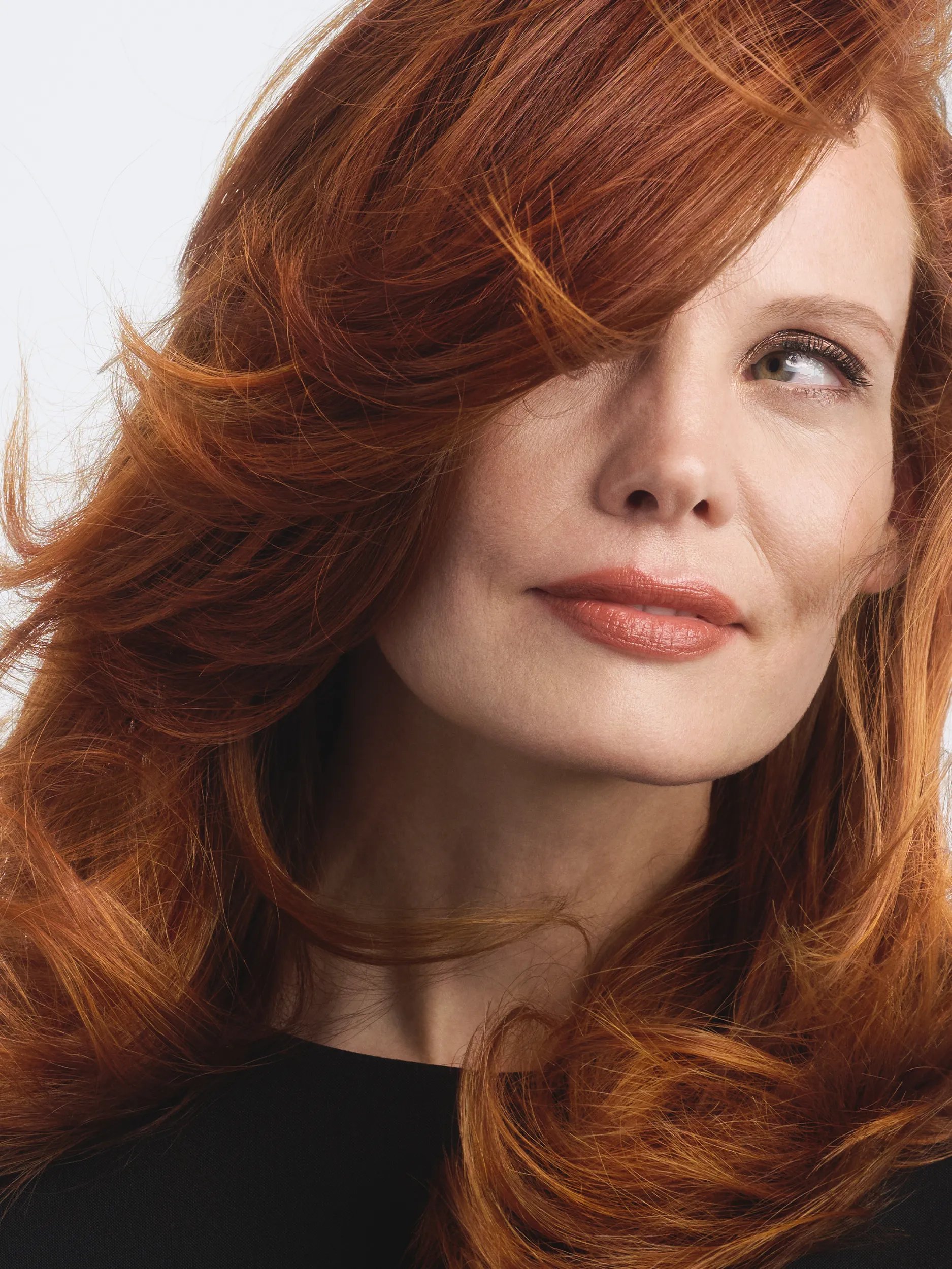 Εικόνα μοντέλου με κόκκινα, κυμματιστά μαλλιά που φοράει ένα μαύρ τοπ. Τα μαλλιά του μοντέλου καλύπτουν το ένα μάτι, ενώ το άλλο μάτι κοιτάει στο πλάι.	