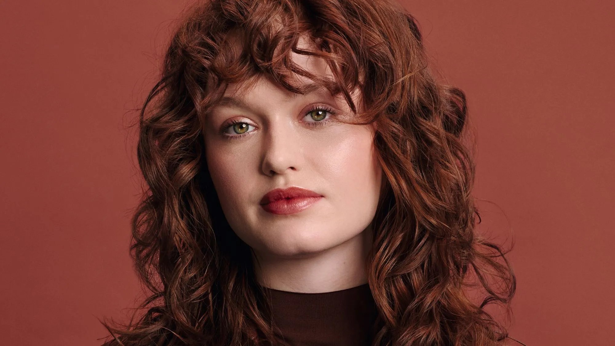 Model with red curly hair, wearing a red leather jacket against a red background. 