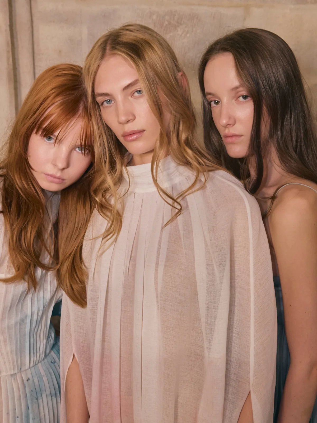 Three models pose backstage – one with copper hair, one with blonde waves, and one with brown, tousled hair.