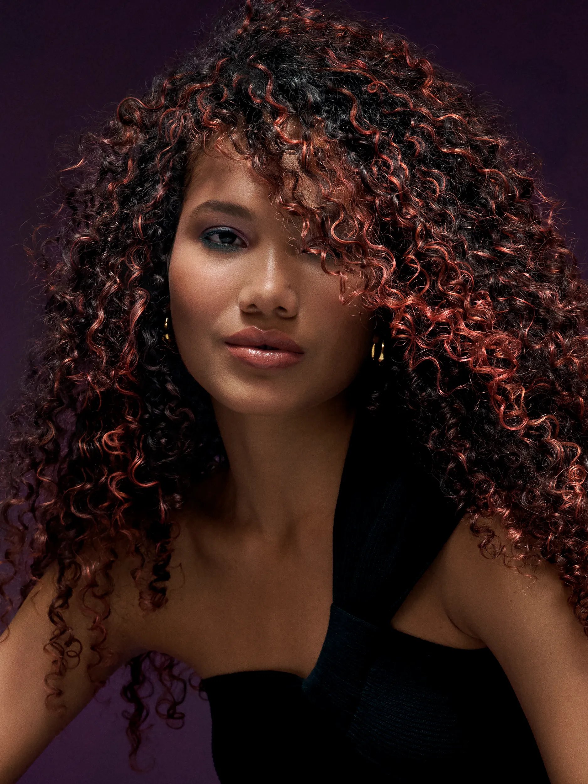 Woman smiling with curly red hair partially covering her face, wearing a sheer red top against a matching background.