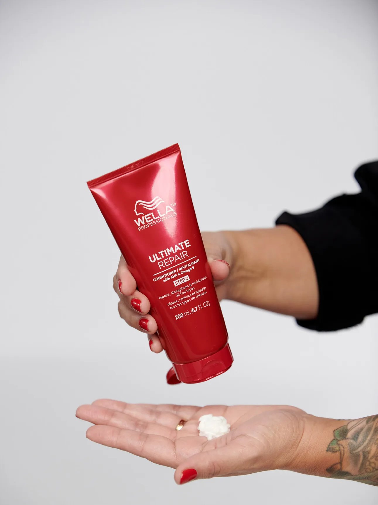 Close-up of hands with red nail polish squeezing Wella Professionals Ultimate Repair conditioner from a red tube into an open palm against a light grey background.