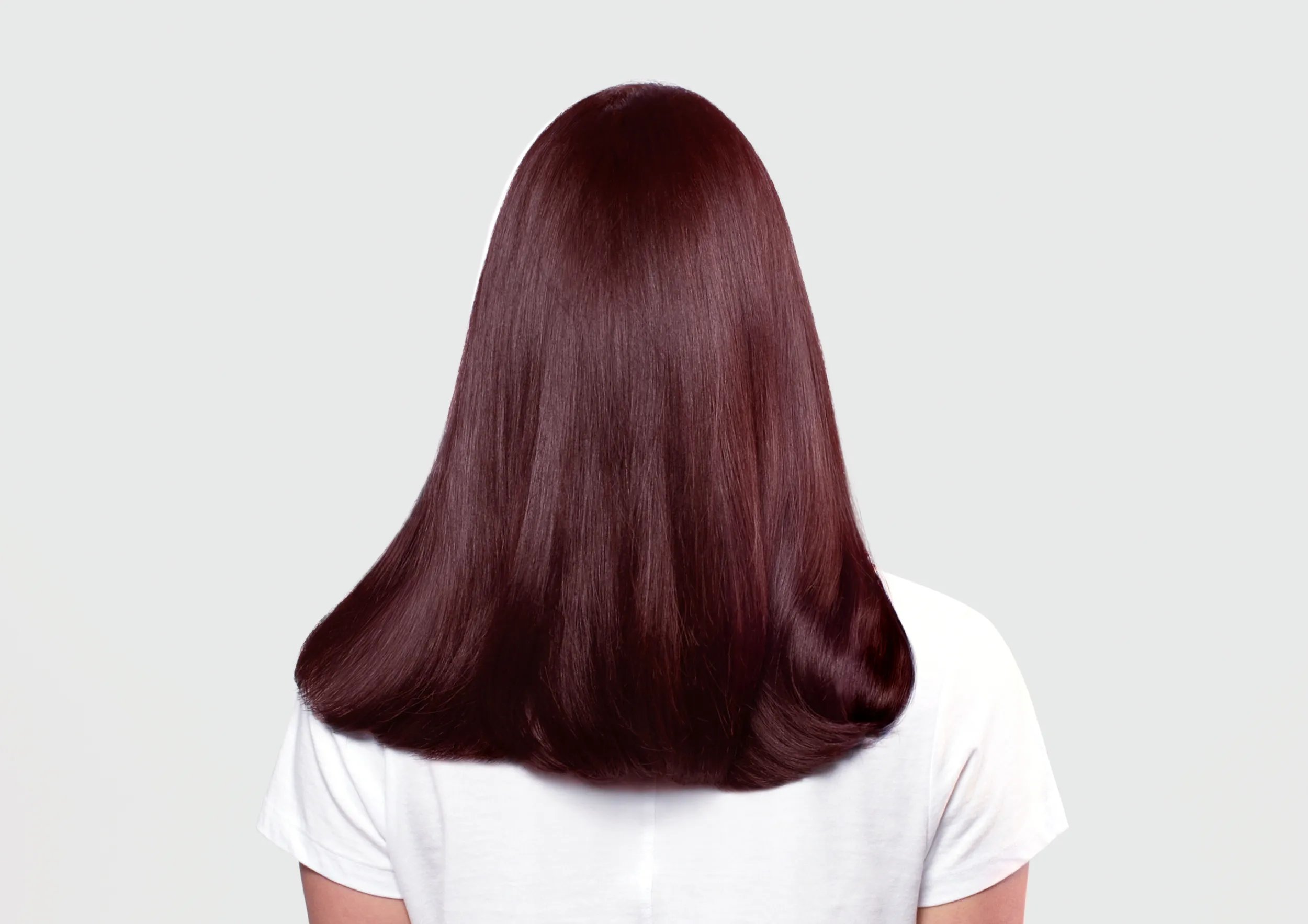 Back view of woman with smooth, shiny burgundy hair wearing a white t-shirt on a light gray background.