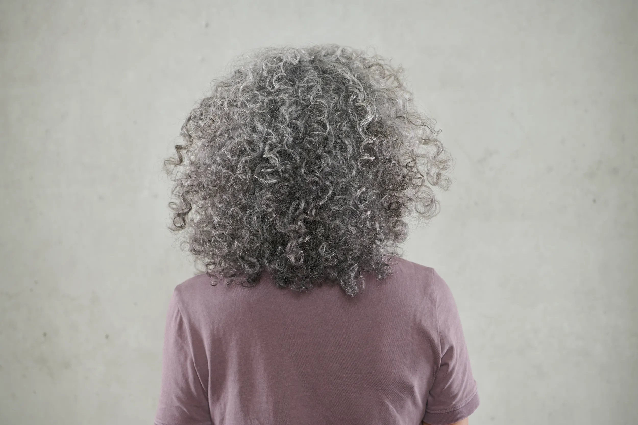 Back of woman's head with curly gray hair on a gray background.