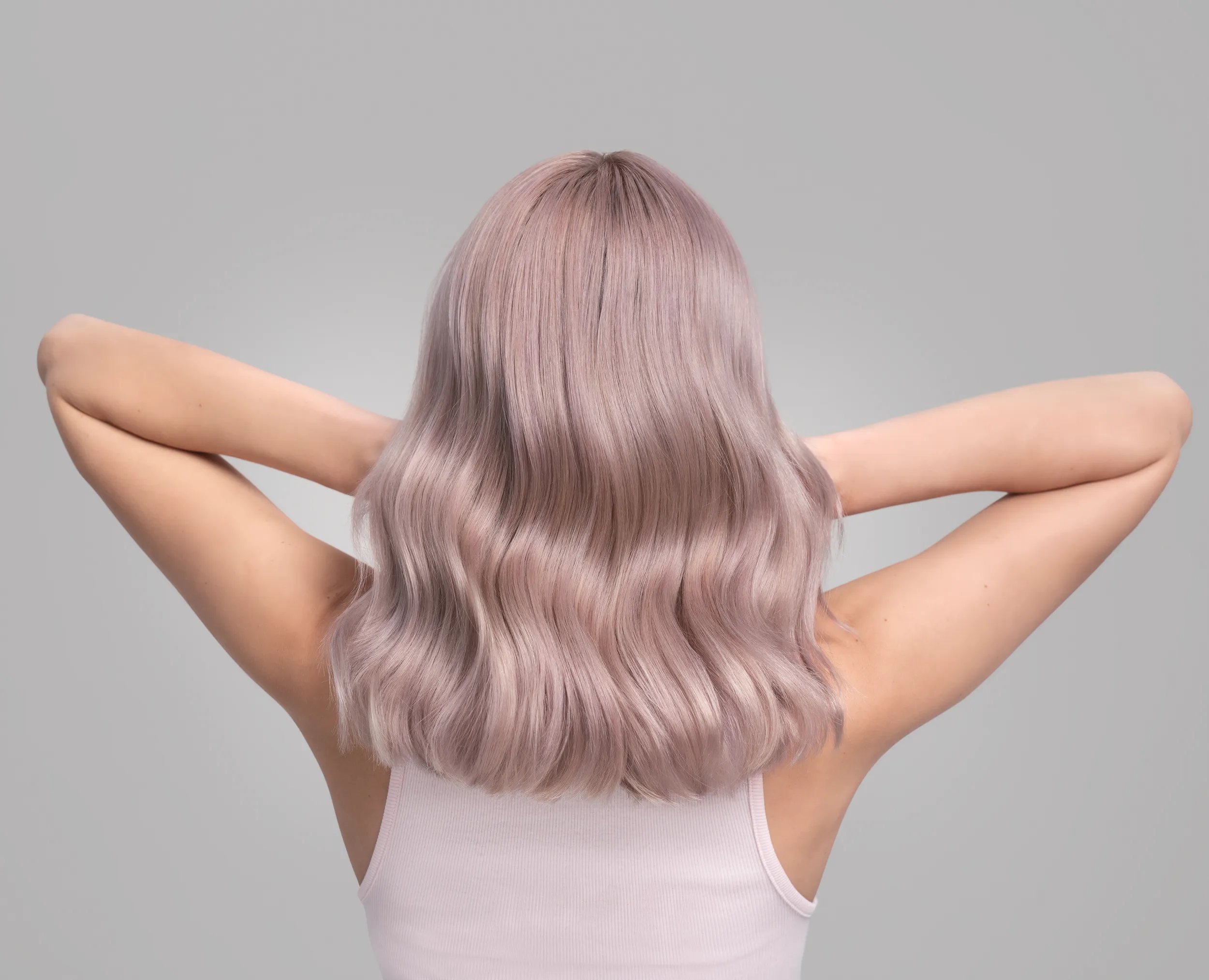 A woman with soft, wavy, pastel lavender hair poses with her hands resting behind her head, wearing a white tank top against a neutral gray background.