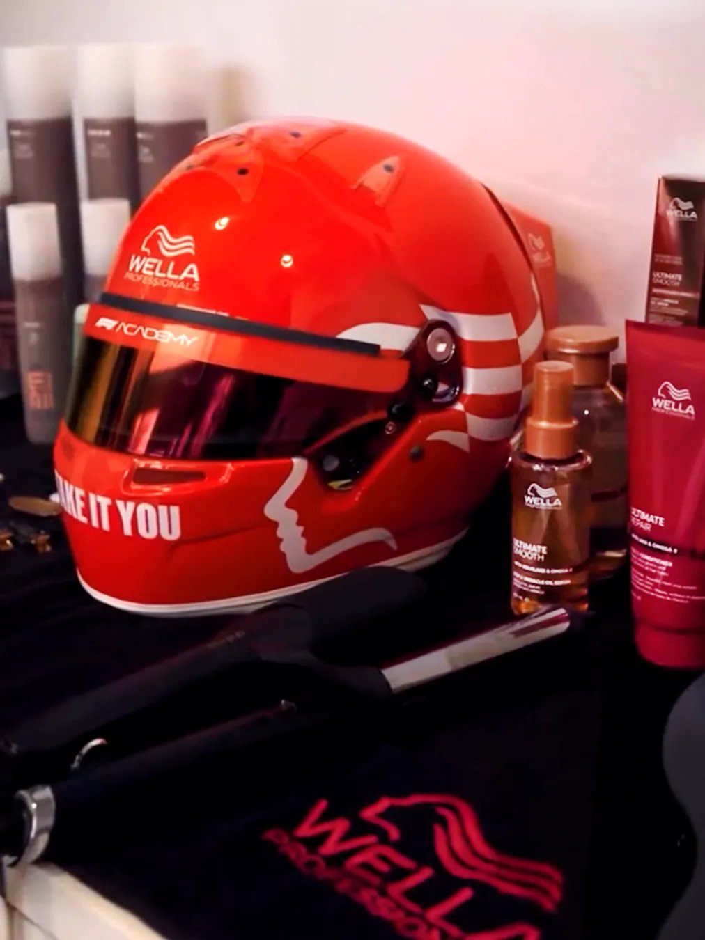 A red racing helmet displayed on a table alongside various Wella haircare and styling products, including bottles and tubes, with a flat iron and a black towel featuring the Wella logo.