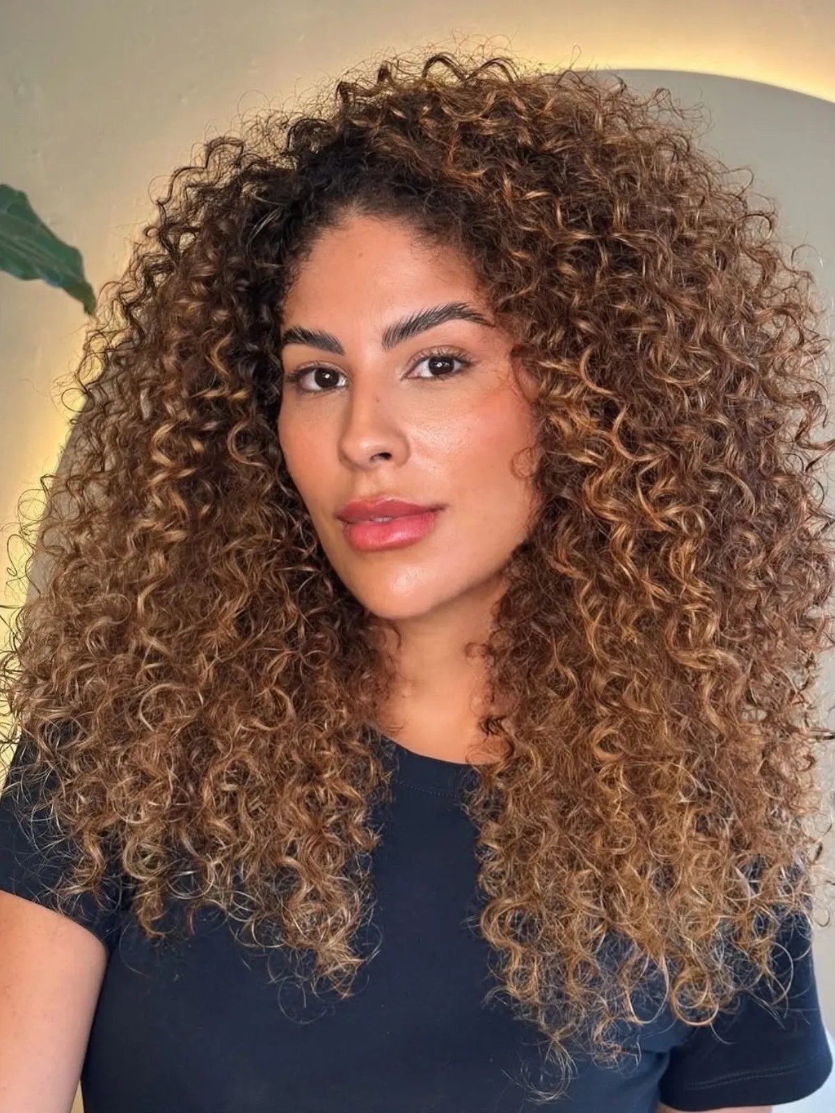 Model with long, glossy, light brown, curly hair, featuring caramel balayage.
