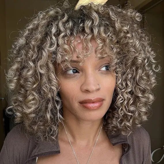 Model with shoulder-length curly hair featuring ash blonde highlights.