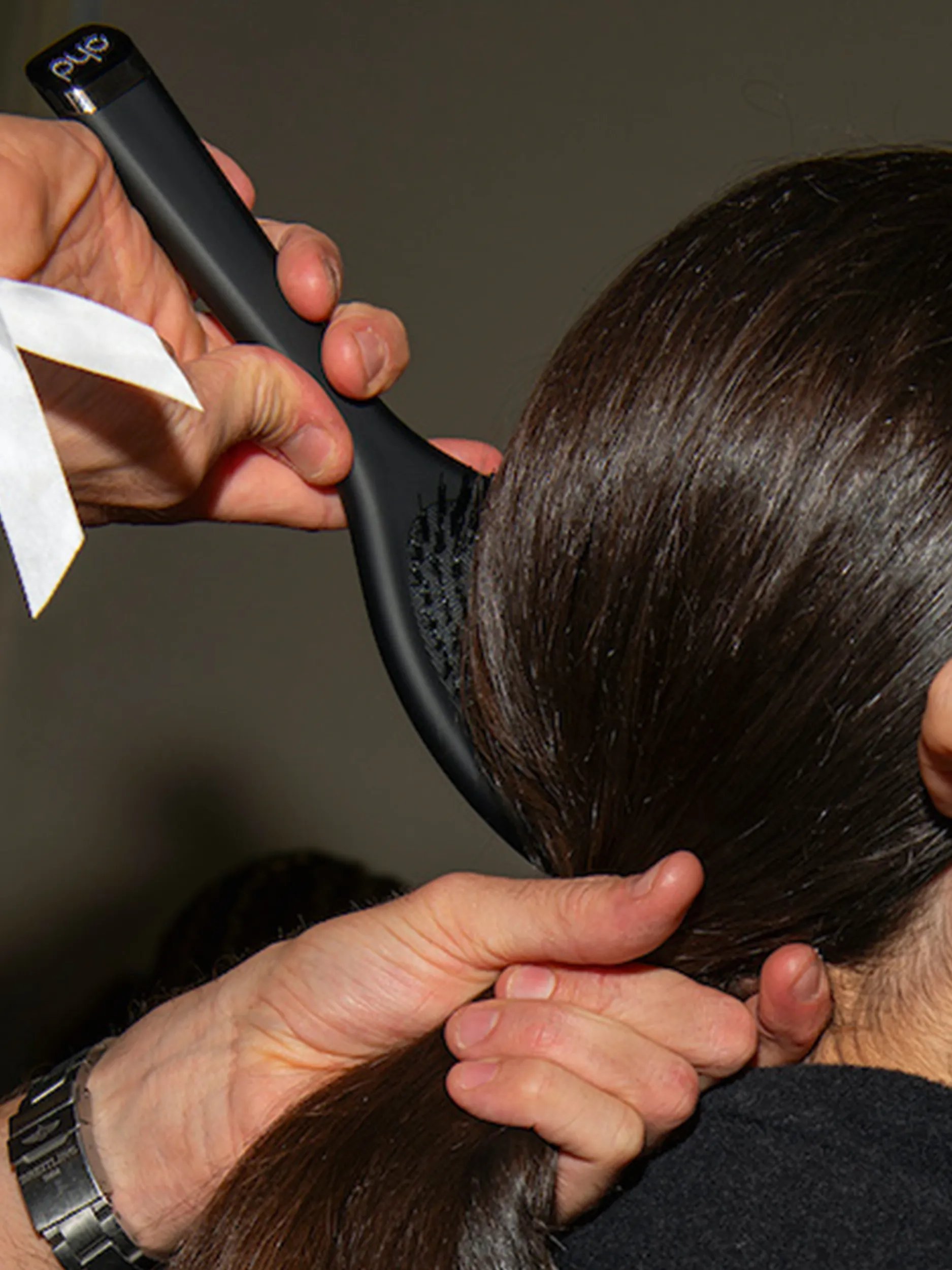 A hairstylist using a black ghd paddle brush to smooth dark brown hair into a ponytail.