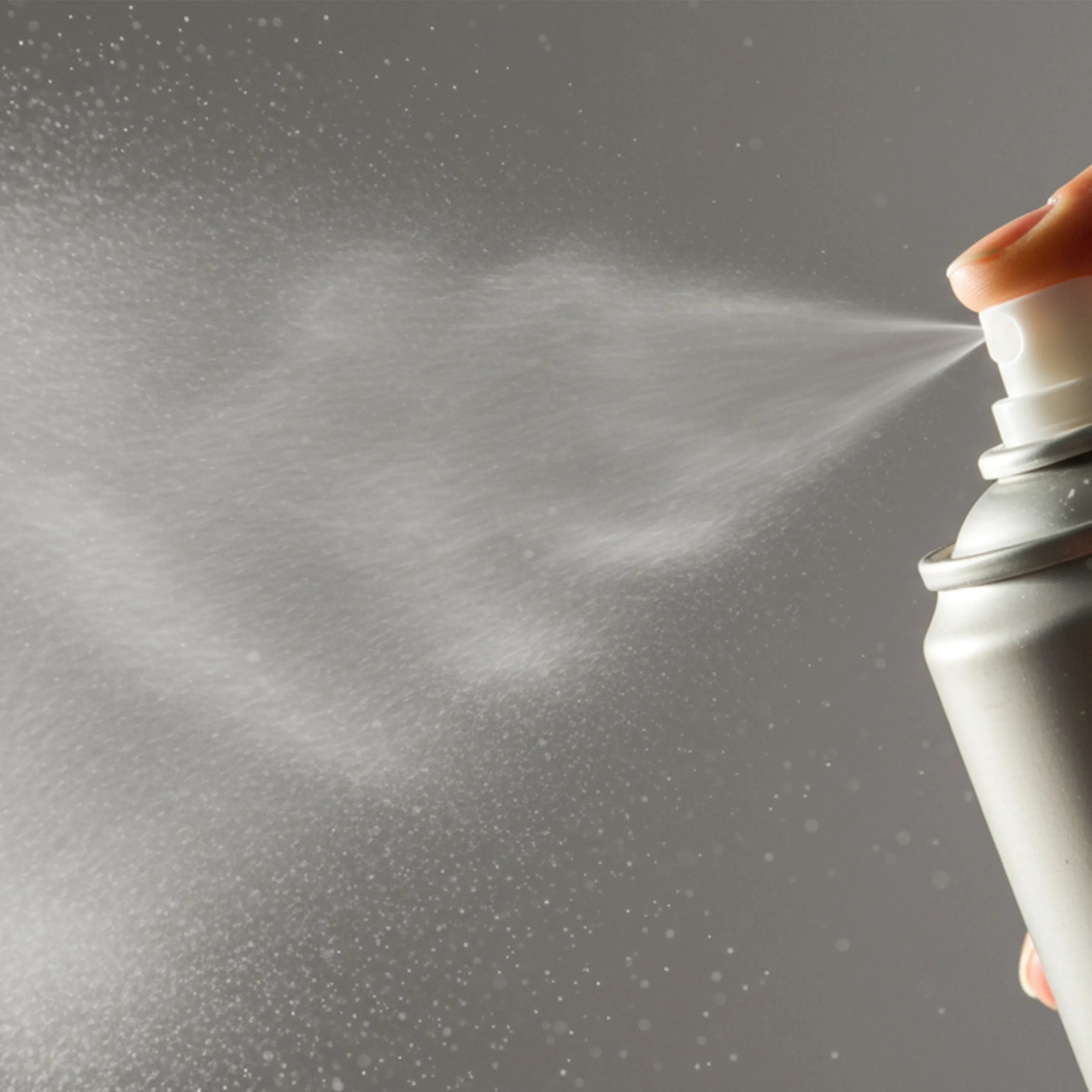 A hand presses the nozzle of an aerosol spray can, releasing a fine mist against a gray background.