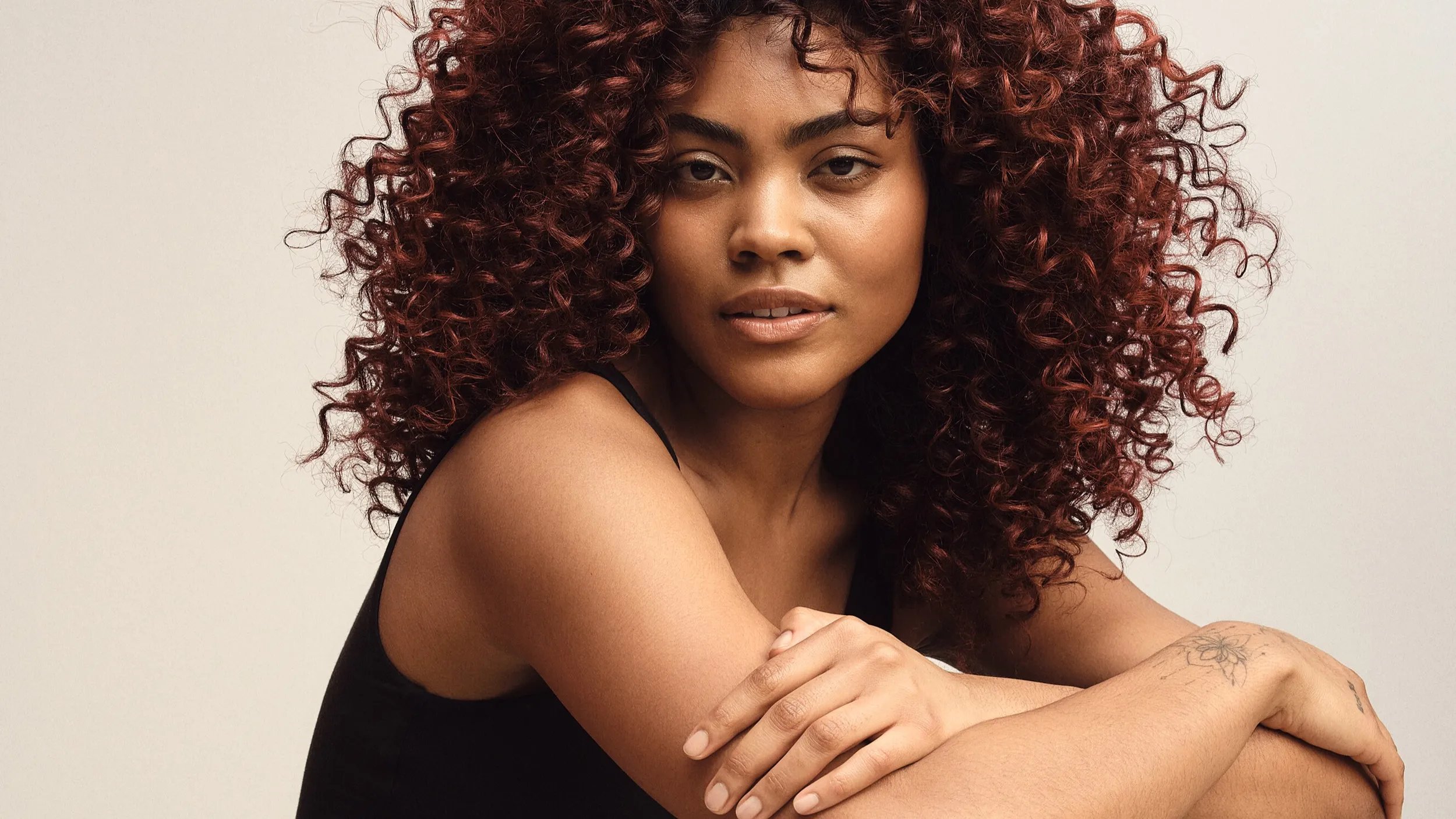 Model with long, curly red hair wearing a sleeveless brown top, looking at the camera against a plain background.