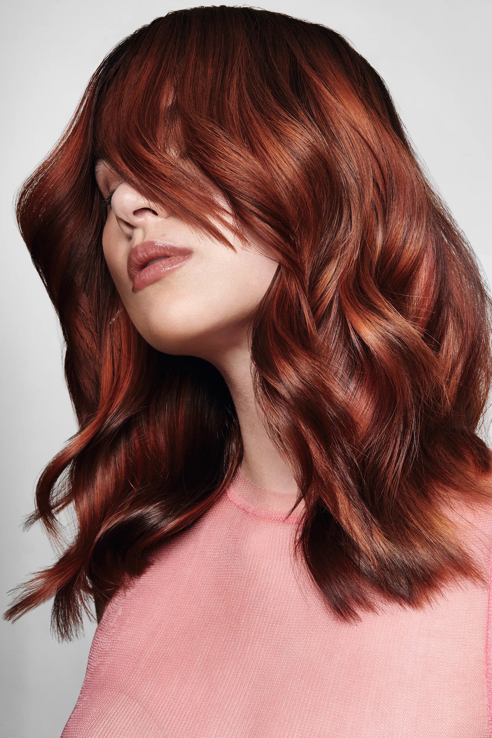 Gros plan d’une femme aux cheveux ondulés couleur auburn coiffés de couches encadrant le visage, portant un haut rose transparent
