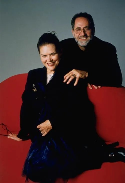 Geri Cusenza and John Sebastian pose together, with Geri seated on a red chair and John standing behind her, both smiling.
