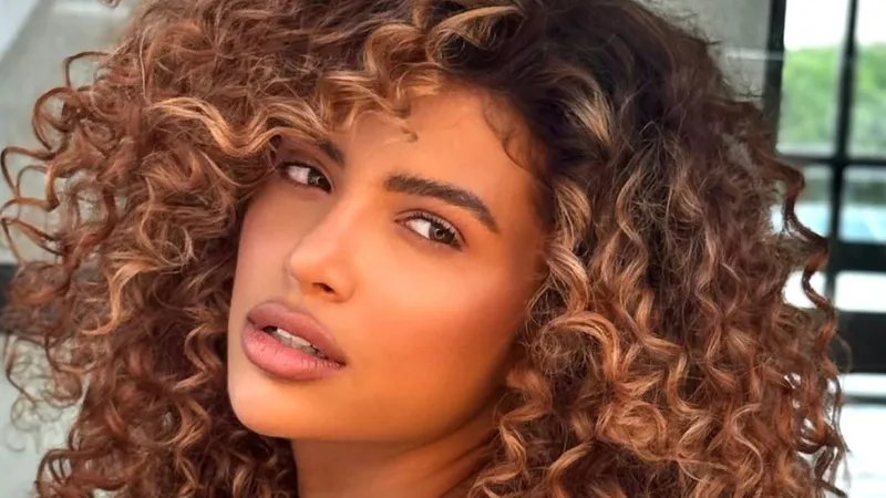 Model with long, dark brown, curly hair featuring golden, sun-kissed highlights.